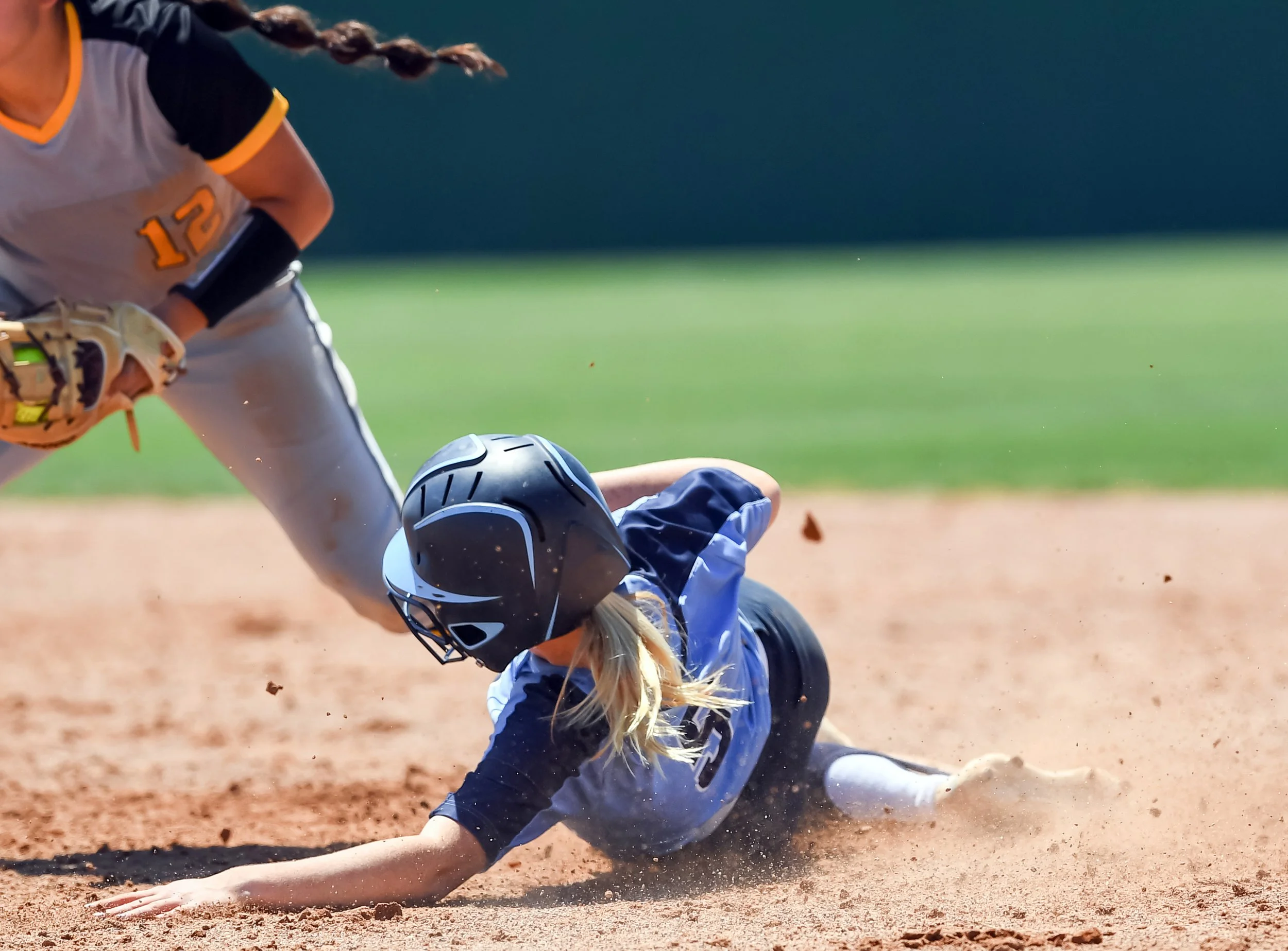 A student-athlete in a blue baseball uniform and helmet is sliding on the dirt during a baseball game, while a player in a white and black uniform with the number 12 is reaching out with a glove to tag them.