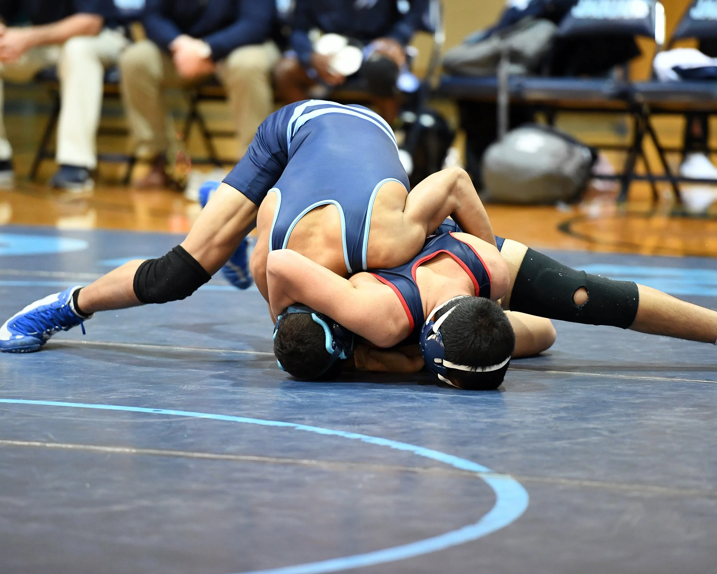 Two wrestlers in a match on a wrestling mat, with one wrestler on top in a pinning position and the other on the bottom, with spectators in the background sitting on benches.