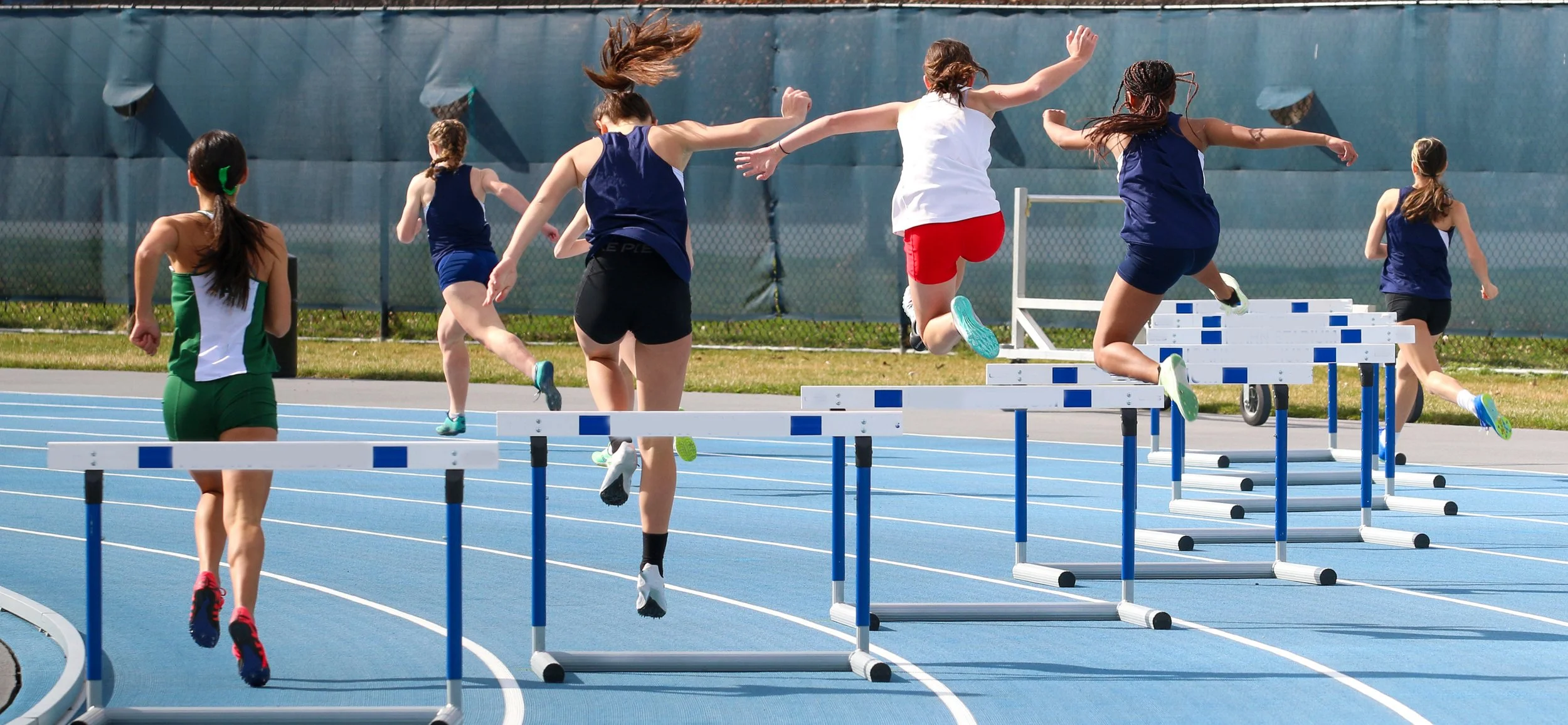 Student-Athletes participating in a hurdles race on a blue running track during daytime.