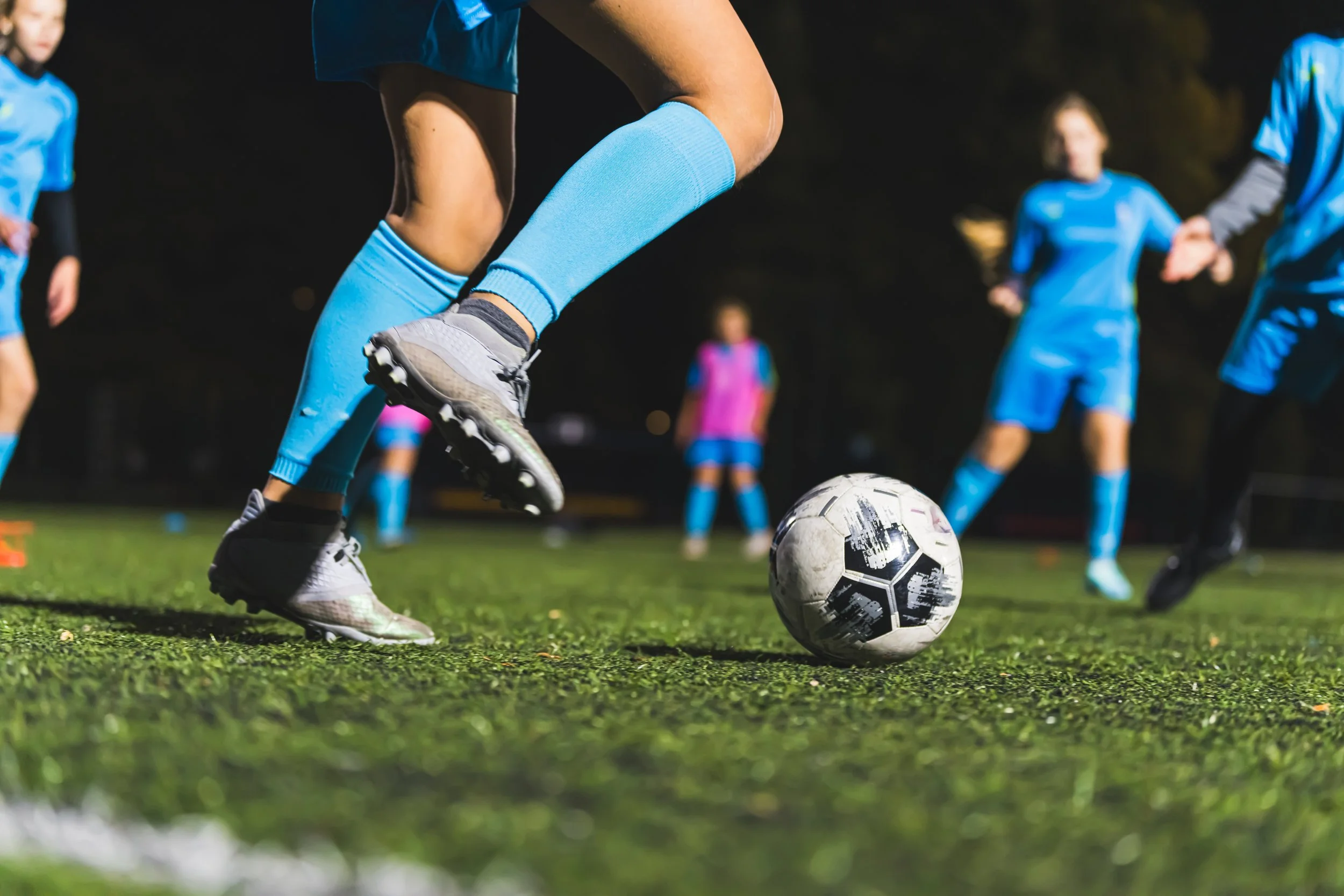 Close-up of a soccer player kicking a soccer ball on a field at night, with other players in blue uniforms in the background.