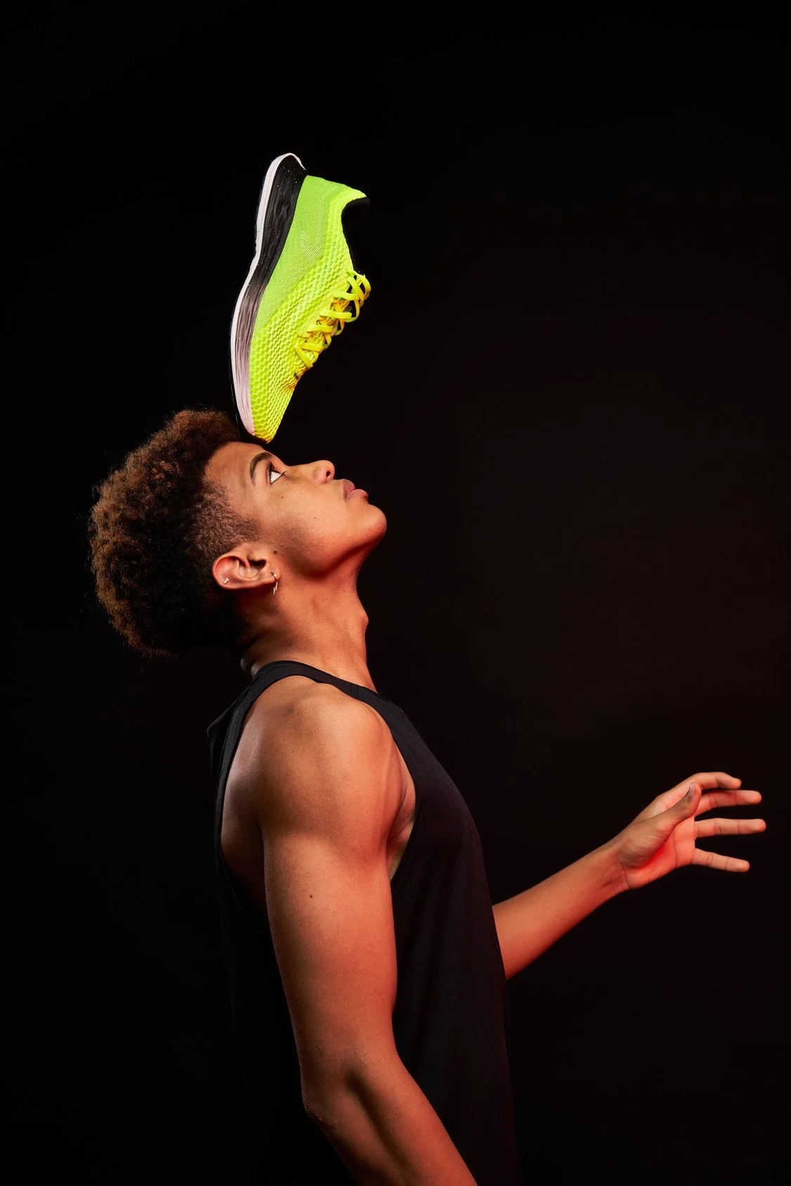 A student-athlete balancing a neon yellow running shoe on their forehead, against a black background.
