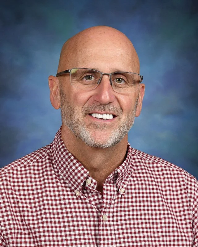 Portrait of a smiling middle-aged man wearing glasses and a red checkered shirt against a blue background.