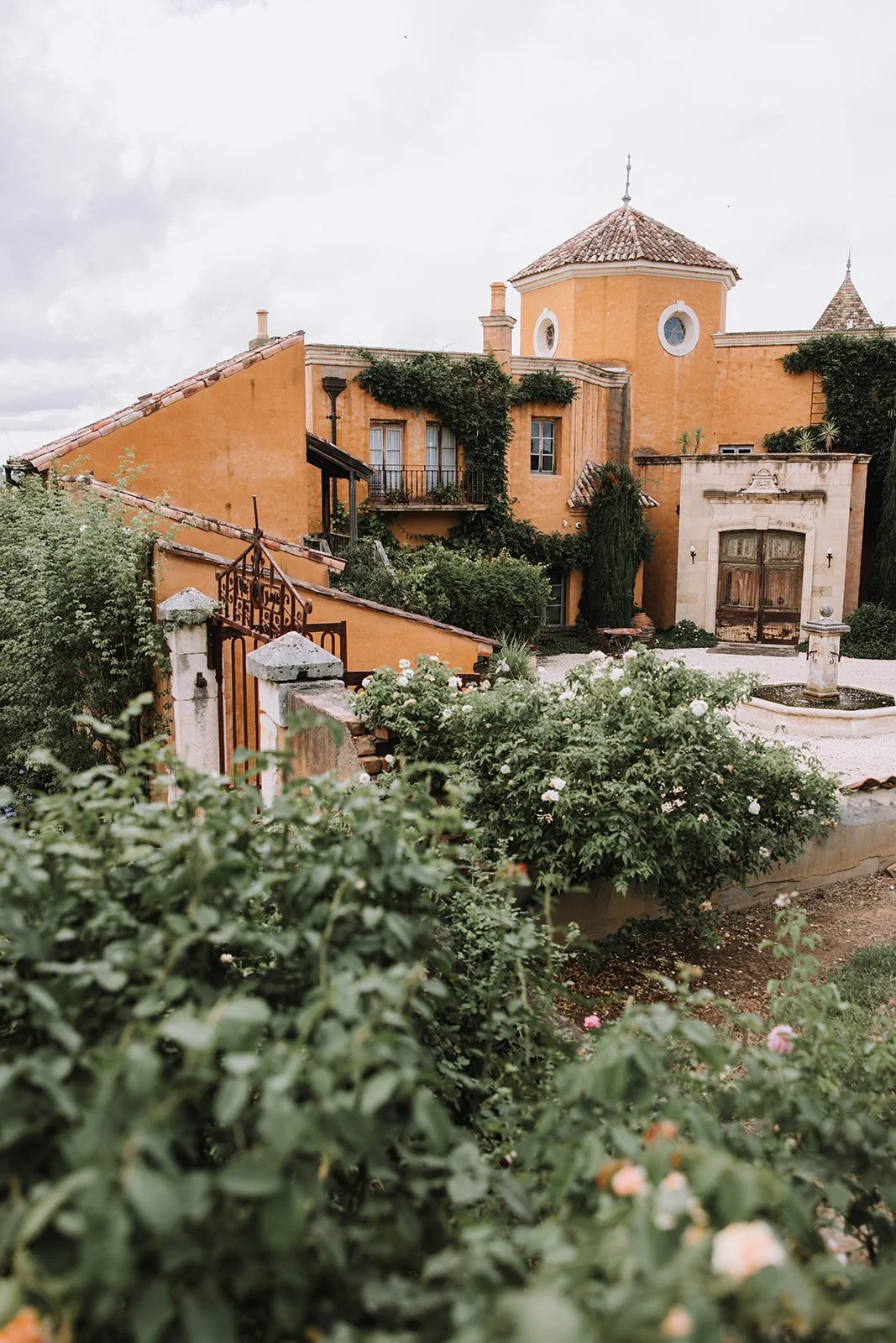 A yellow Mediterranean-style house with a red-tiled roof, surrounded by lush green bushes and blooming flowers, with an ornate wooden door and a tower-like structure on the right.