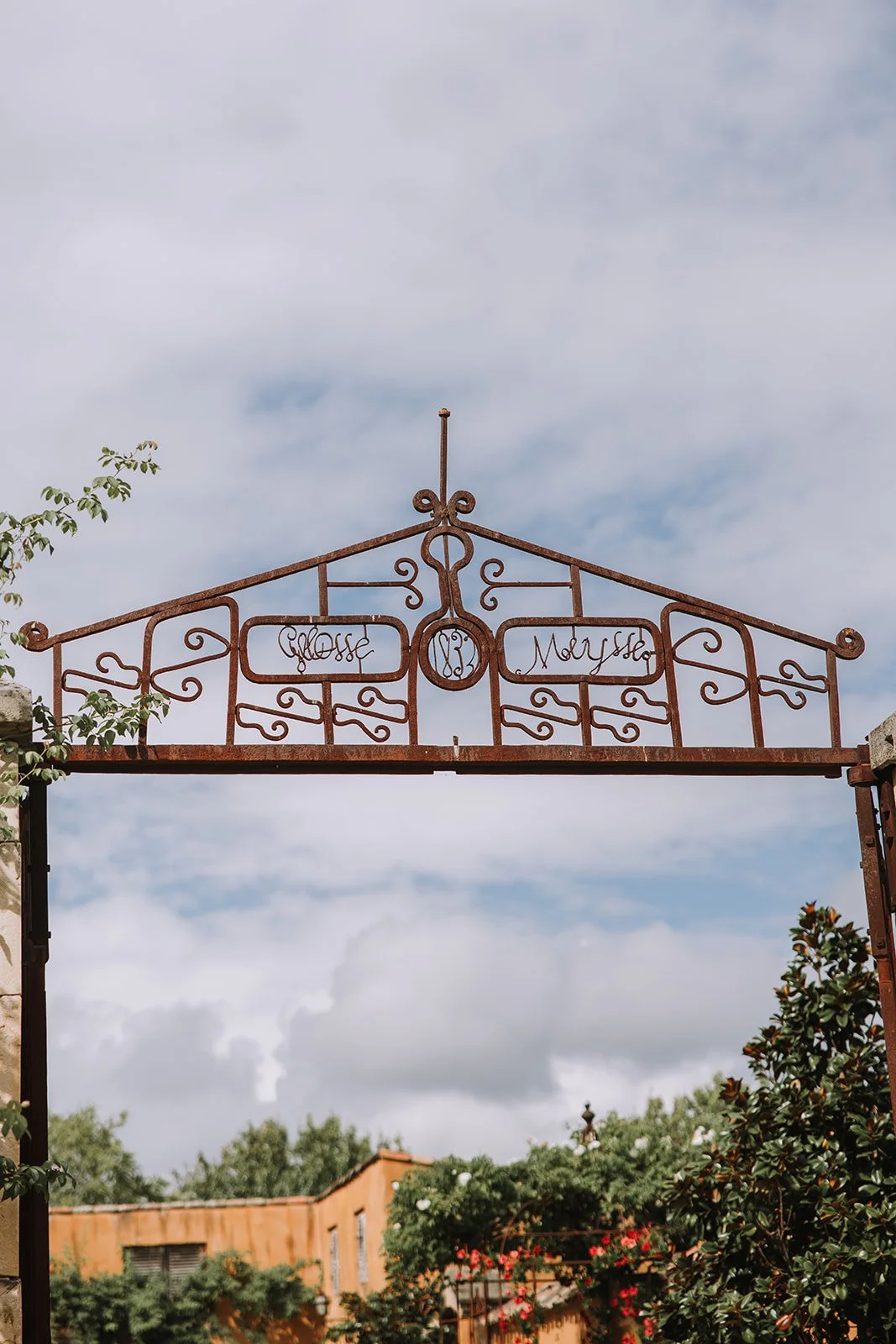 Rusty metal archway with decorative scrolling and writing, set against a cloudy sky, with trees and a building in the background.