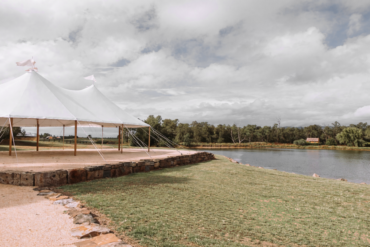 A large white outdoor tent with flags on top, set on a stone platform near a body of water, with trees and cloudy sky in the background.