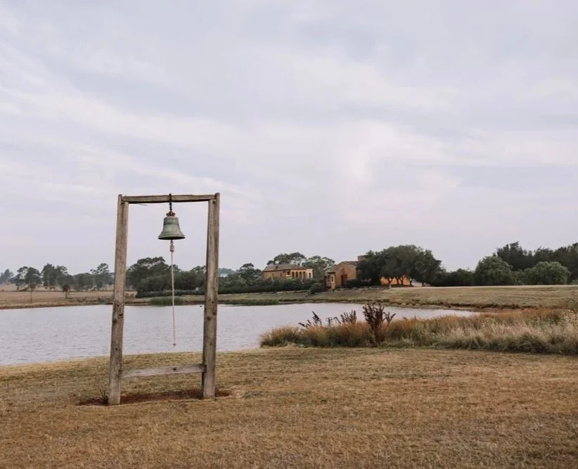 A large outdoor bell hanging from a wooden frame near a lake with trees and buildings in the background.