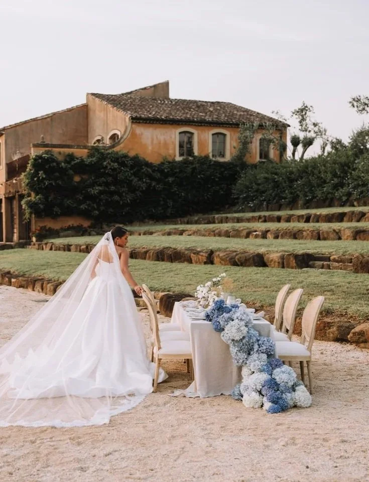 Bride in a white wedding dress with a long veil setting a formal table outdoors with a house and greenery in the background.