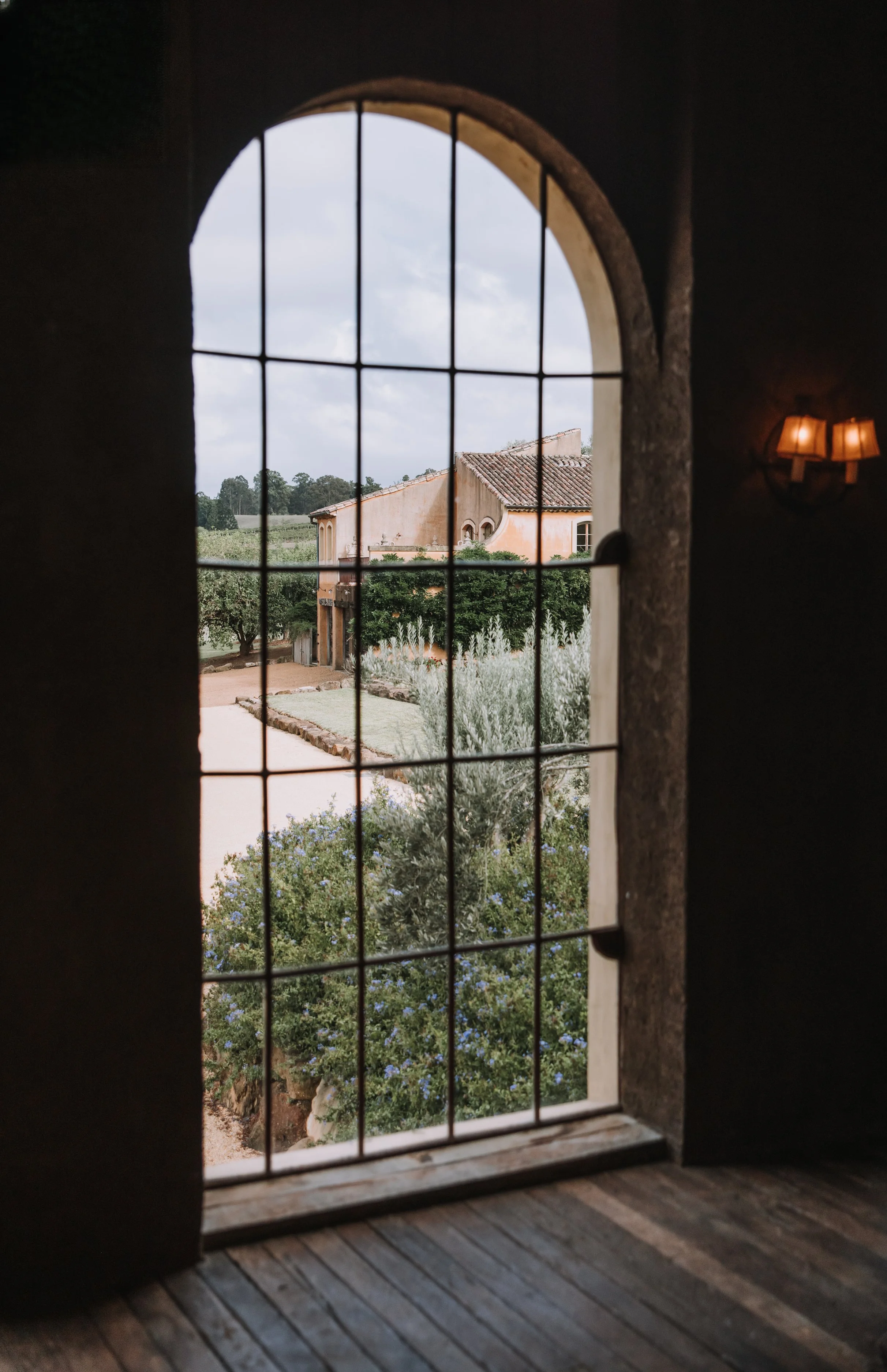 View through a grid window showing a courtyard with green trees, a dirt pathway, and a beige stucco building with a tiled roof under a cloudy sky.
