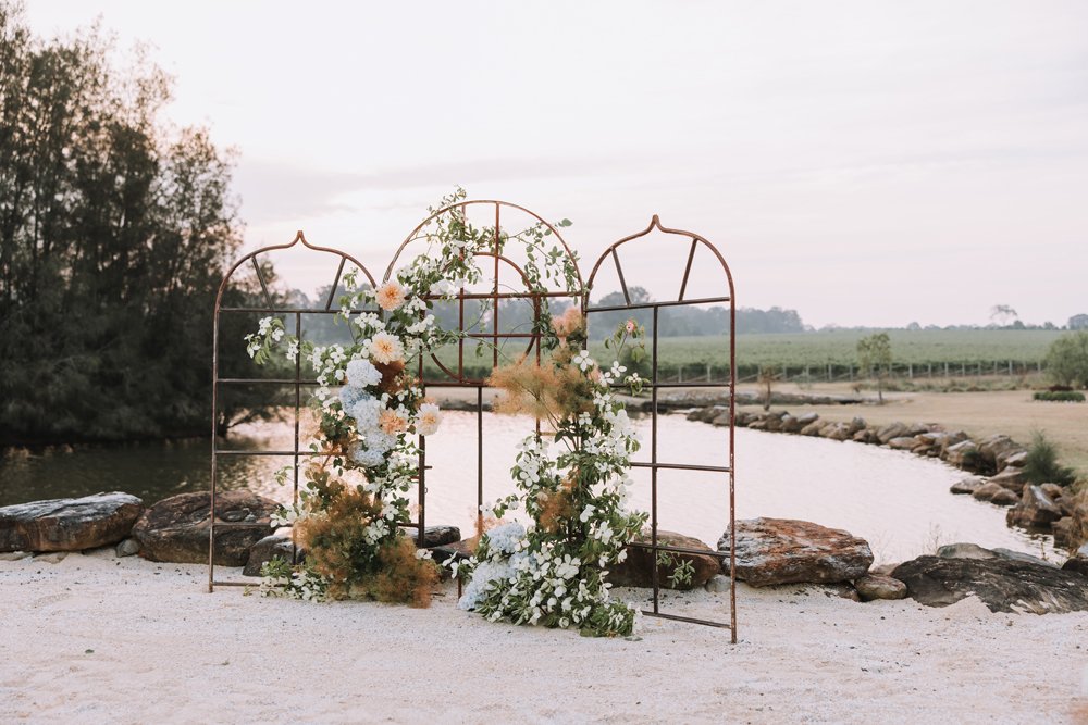 A floral wedding arch with white and pastel flowers and greenery, set on a sandy surface near a river with rocks, trees, and fields in the background during sunset.