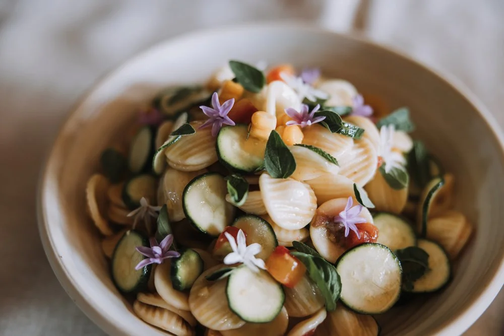 A bowl of pasta salad with zucchini, basil, and purple edible flowers.