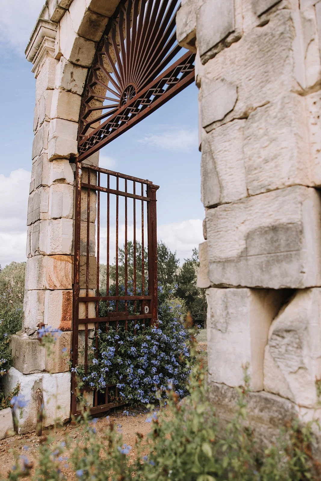 A stone archway with an iron gate and decorative fan-shaped ironwork above it. There are purple flowers at the base of the gate and trees in the background.