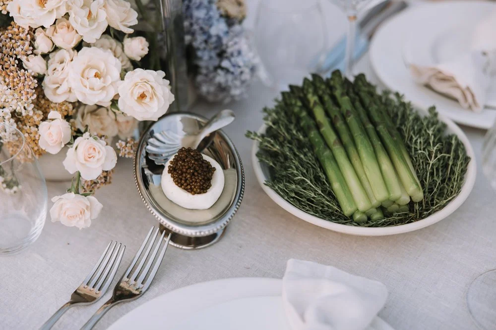 A table setting with a plate, two forks, a small bowl with a  black caviar, a large platter of asparagus, and a bouquet of light pink and white flowers.