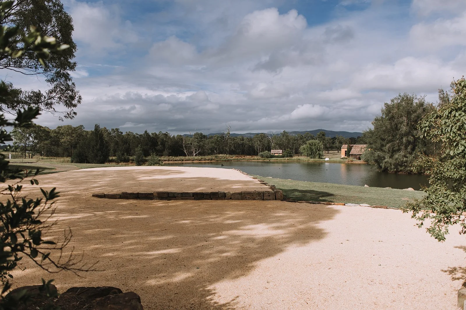 A scenic landscape featuring a sandy area in the foreground, a pond or small lake on the right, surrounded by trees and greenery. There are hills or mountains in the distance under a partly cloudy sky.