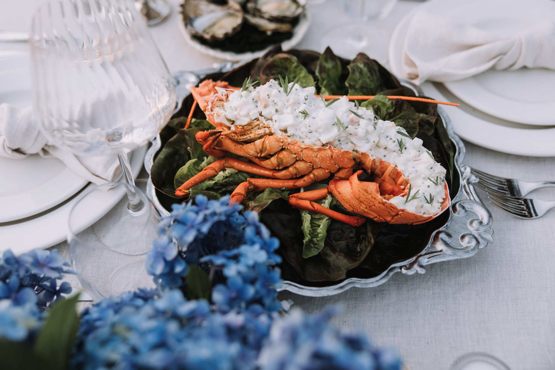 A seafood dish featuring lobster, topped with a creamy salad and garnished with herbs, on a silver platter. The table is set with white plates, cutlery, wine glasses, and napkins, and decorated with blue flowers.