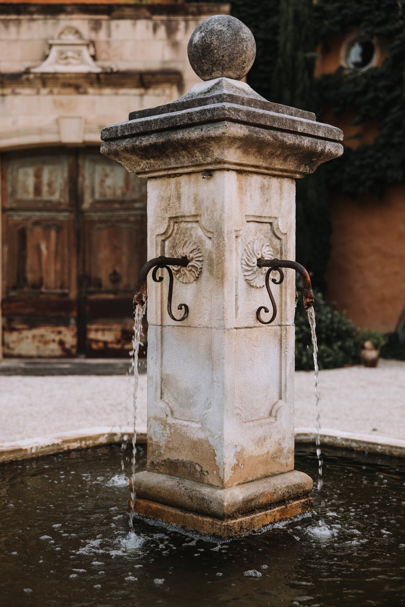 Stone fountain with water flowing from two spouts, situated in an outdoor garden area.