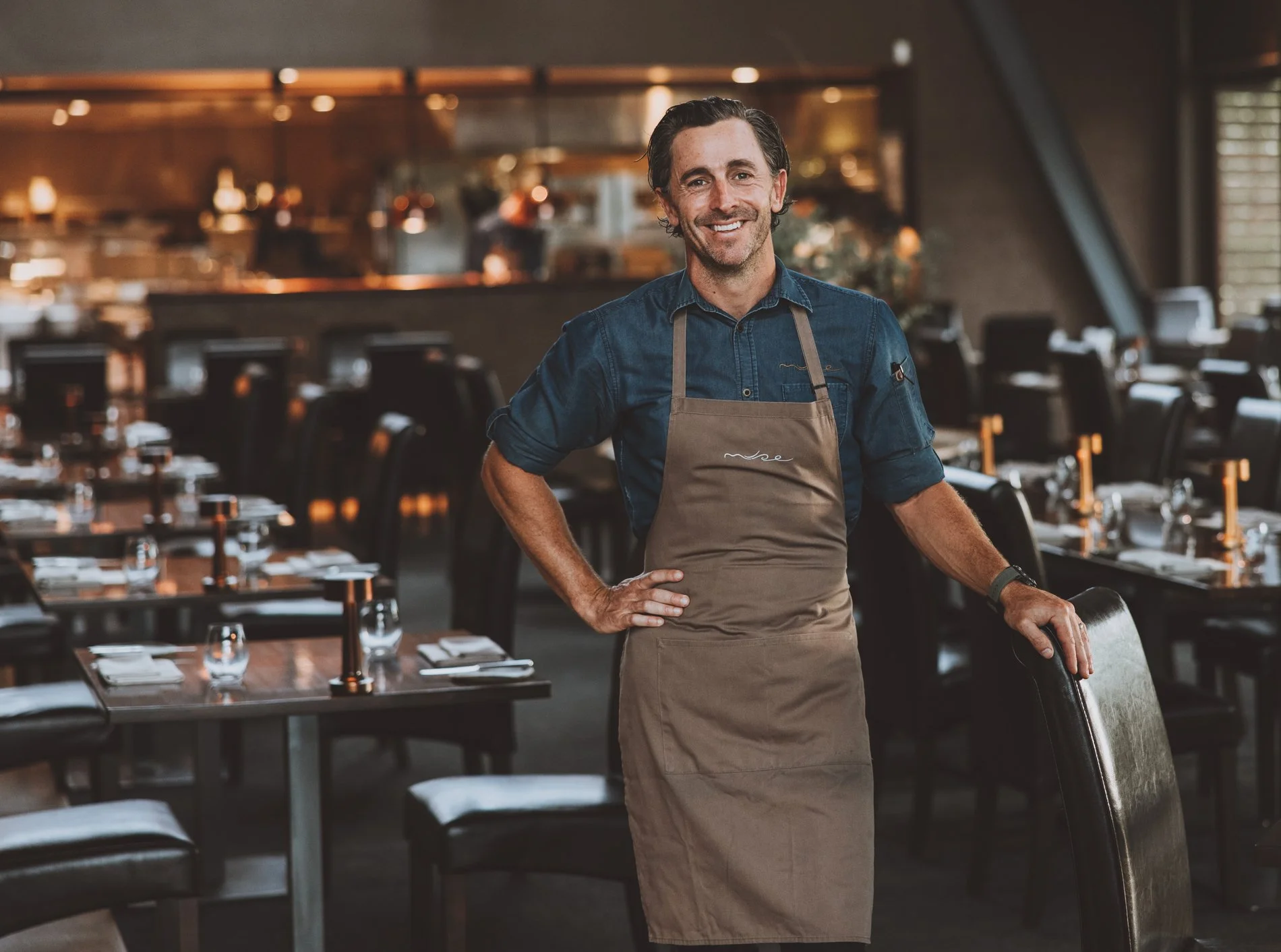 Troy Rhoades-Brown from Muse Restaurant wearing a blue Chef jacket and a brown apron standing in Muse restaurant with tables set for dining.