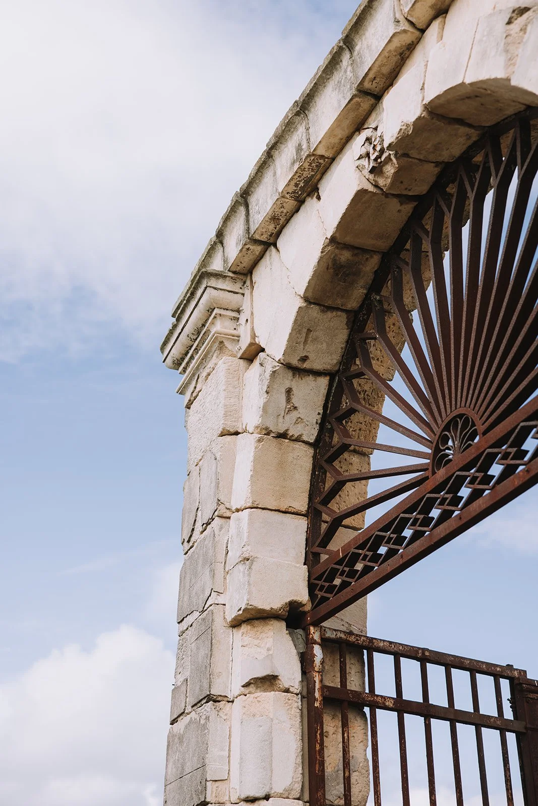 Close-up of an old stone archway with a decorative wrought iron gate, against a blue sky with scattered clouds.