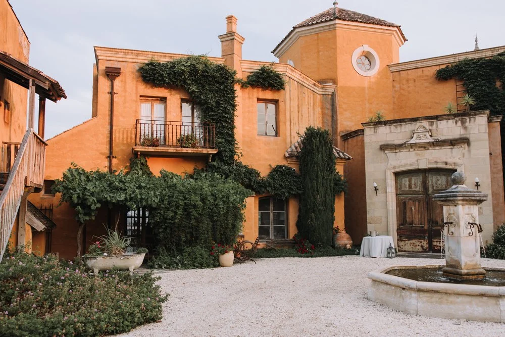 A European-inspired castle, a small tower with a round window, greenery climbing the walls, a courtyard with a stone fountain, potted plants.