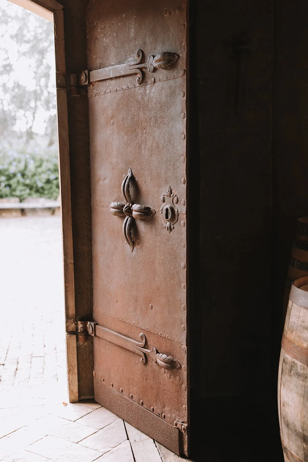 An old, rusty iron door with decorative leaf-shaped handles partially open, revealing a dark interior, with a stone or brick floor and barrels to the side.