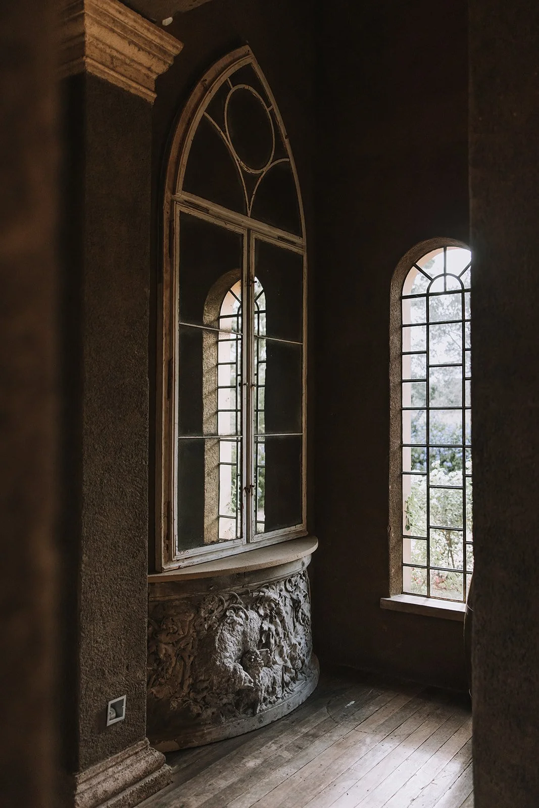 Interior of an old building with tall arched windows, one with a decorative wooden frame, a textured stone wall feature, and wooden flooring. Light filters through the windows.
