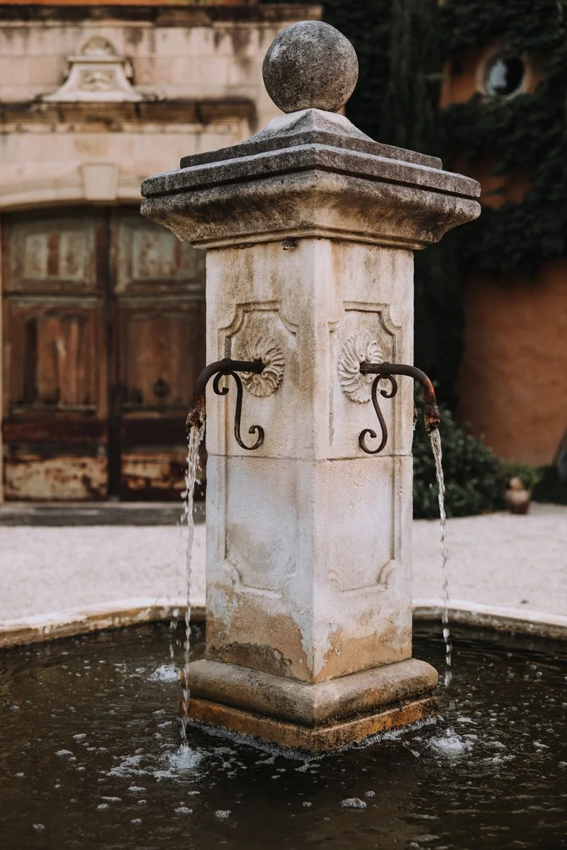 Stone fountain with water spouting from two metal spouts, surrounded by a shallow pool, with a historical building and greenery in the background.