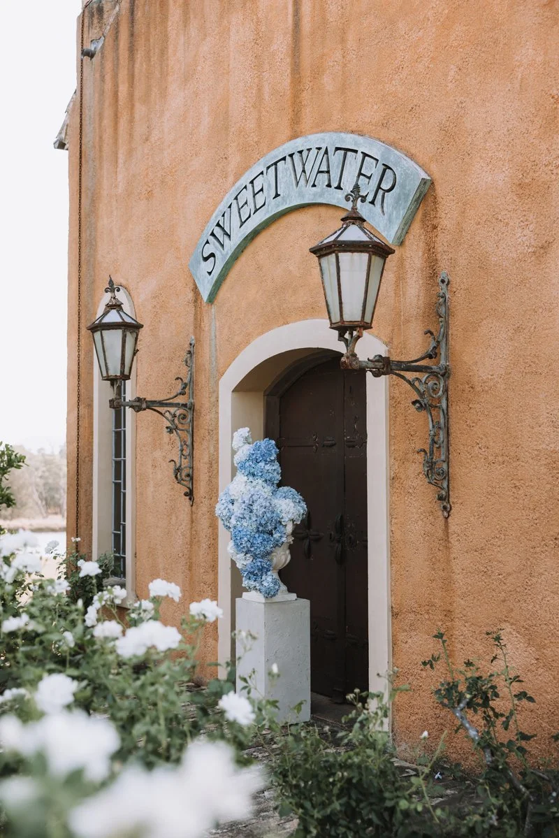 A building with an European exterior, a dark wooden door, and a sign above that reads 'Sweetwater'. There are two ornate lantern-style light fixtures on either side of the door and an urn covered in blue and white flowers in front of the door.