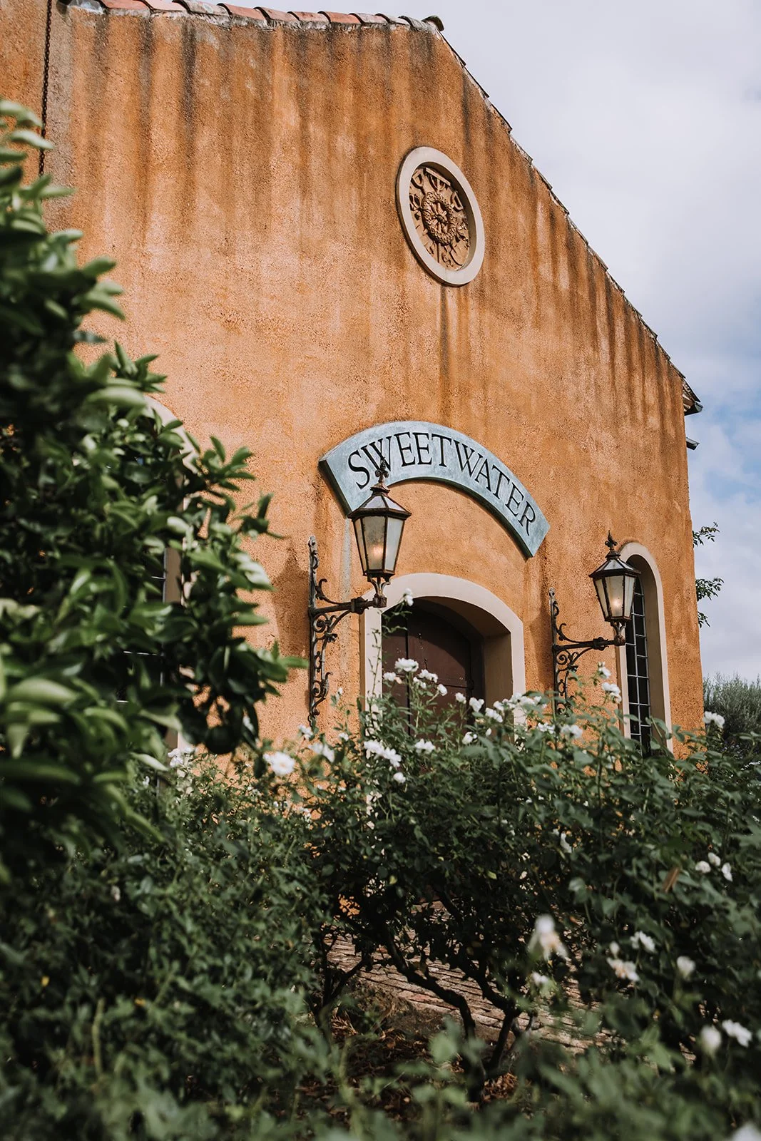 The image shows the exterior of a building with a sign that reads 'Sweetwater'. The building has an orange stucco facade with black wrought iron lamps on either side of an arched dark door. There are white flowering bushes in foreground and a cloudy 