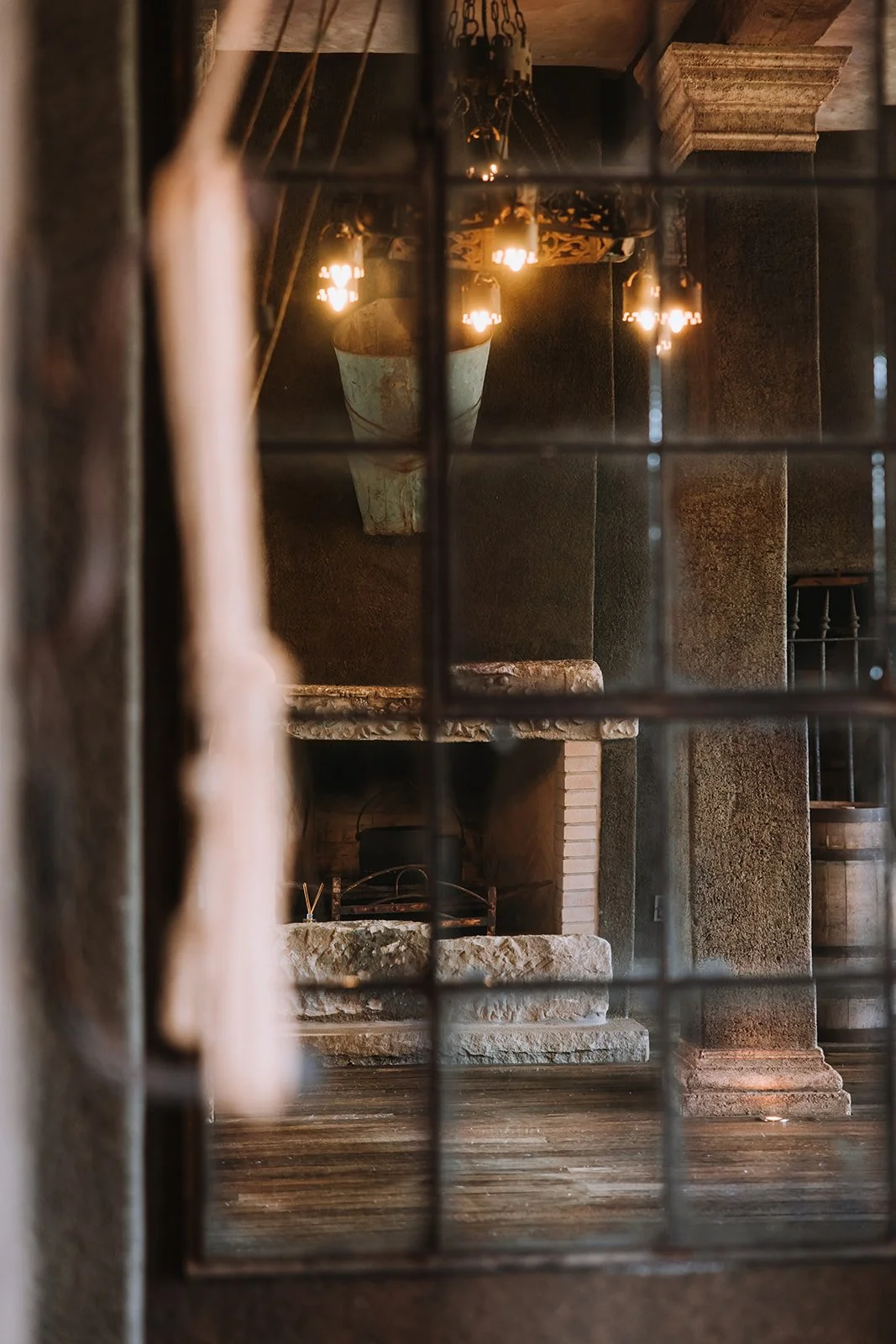 Interior of a rustic room viewed through window bars, featuring a fireplace with a stone mantel, hanging lamps, and wood flooring.