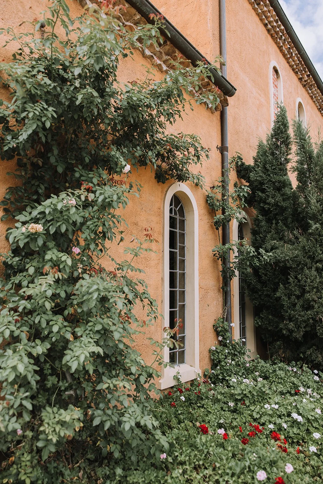 A peach-colored building with tall, narrow arched windows, surrounded by lush green bushes and flowers, with a black rain gutter along the roof edge.