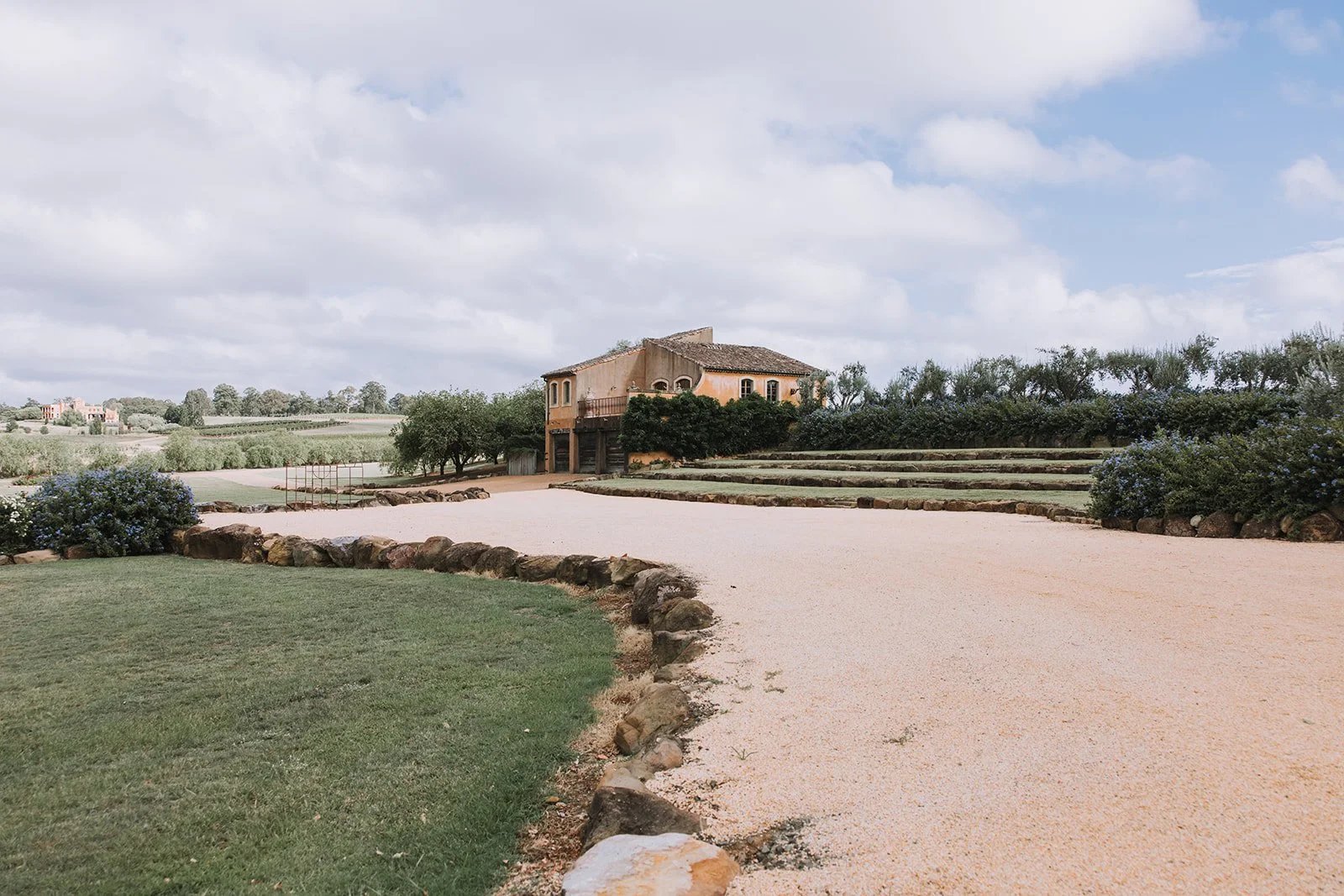 A house in a rural landscape with a gravel pathway, lawn, bushes, and trees under a partly cloudy sky.