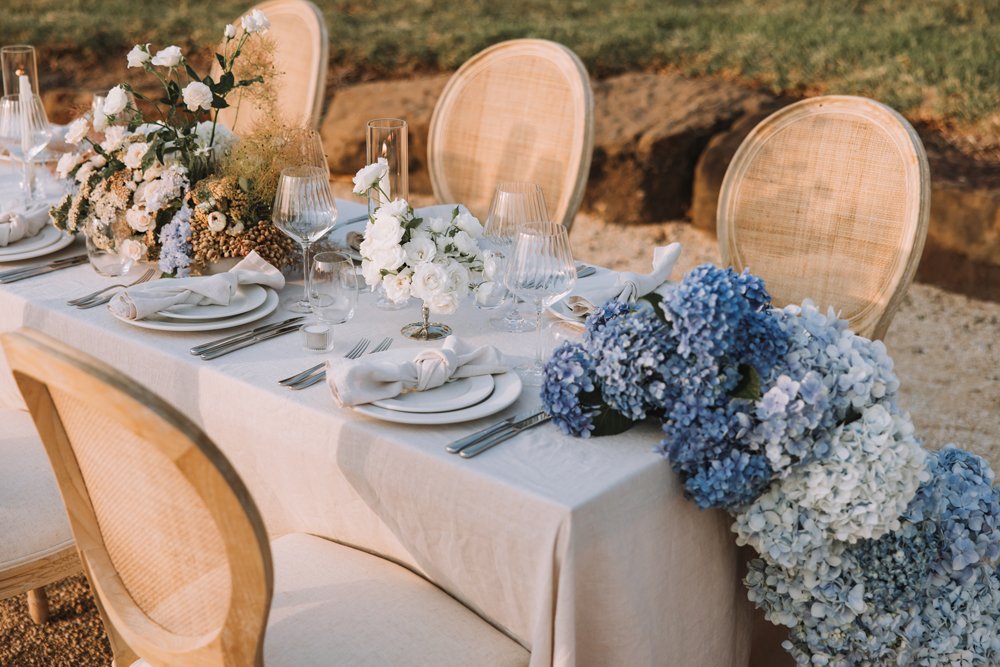 Elegant outdoor dining table decorated with white and blue floral arrangements, set with white plates, napkins, and glassware, with wedding chairs around it.
