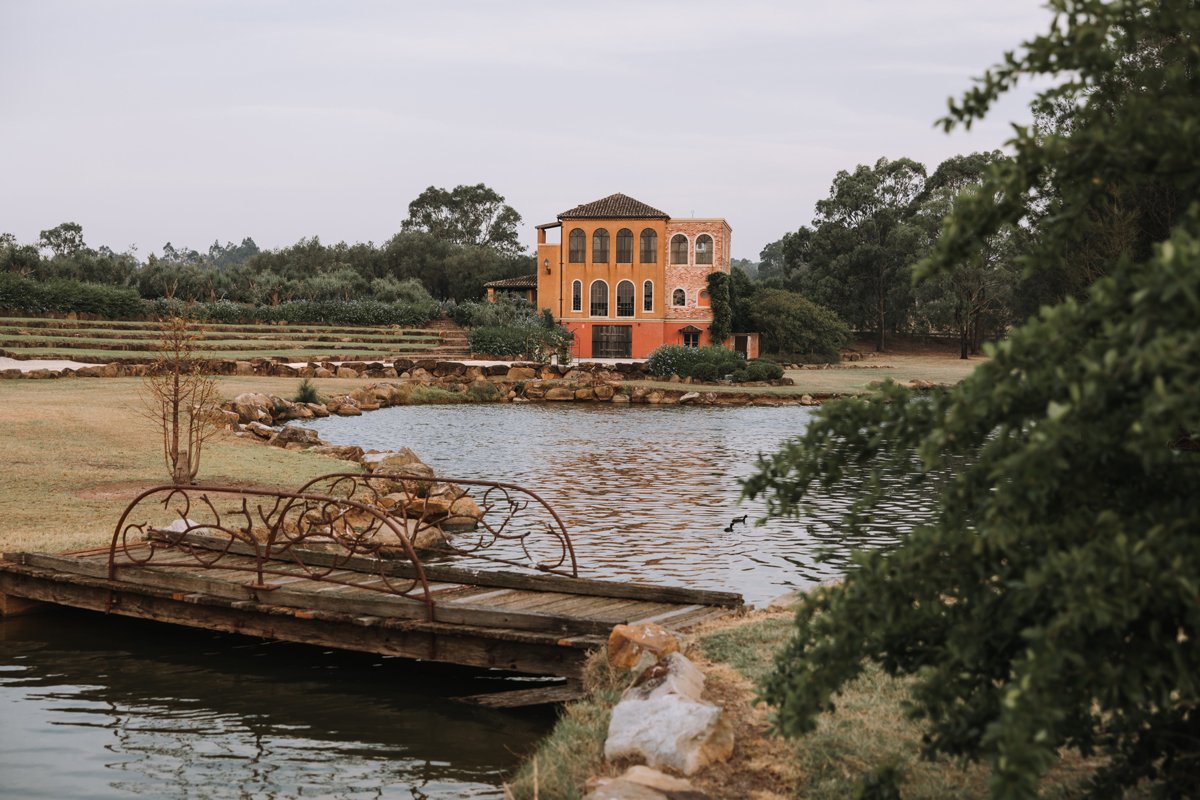 A pond with a small wooden and iron frame bridge in the foreground, a large building with arched windows in the background, surrounded by trees and landscape.