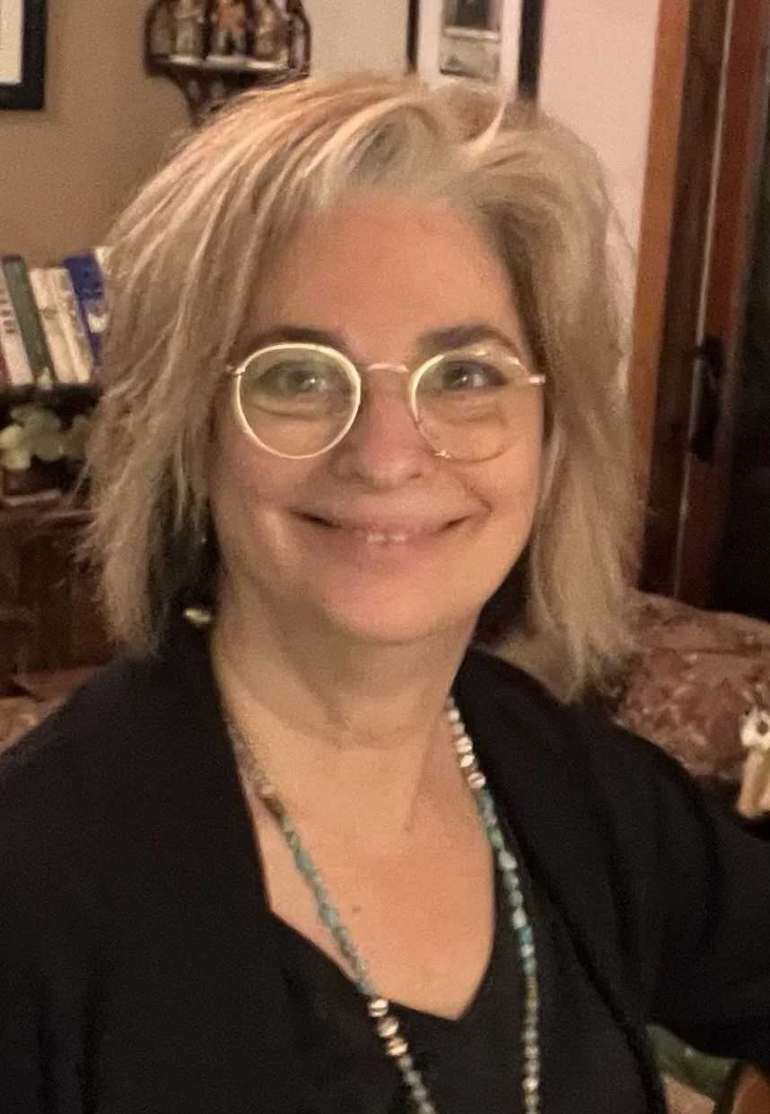 Karen Whalen, founder of Lake Geneva Art Studio | A smiling middle-aged woman with blonde, tousled hair wearing glasses, a black top, and layered necklaces, sitting in a cozy room with bookshelves and framed pictures