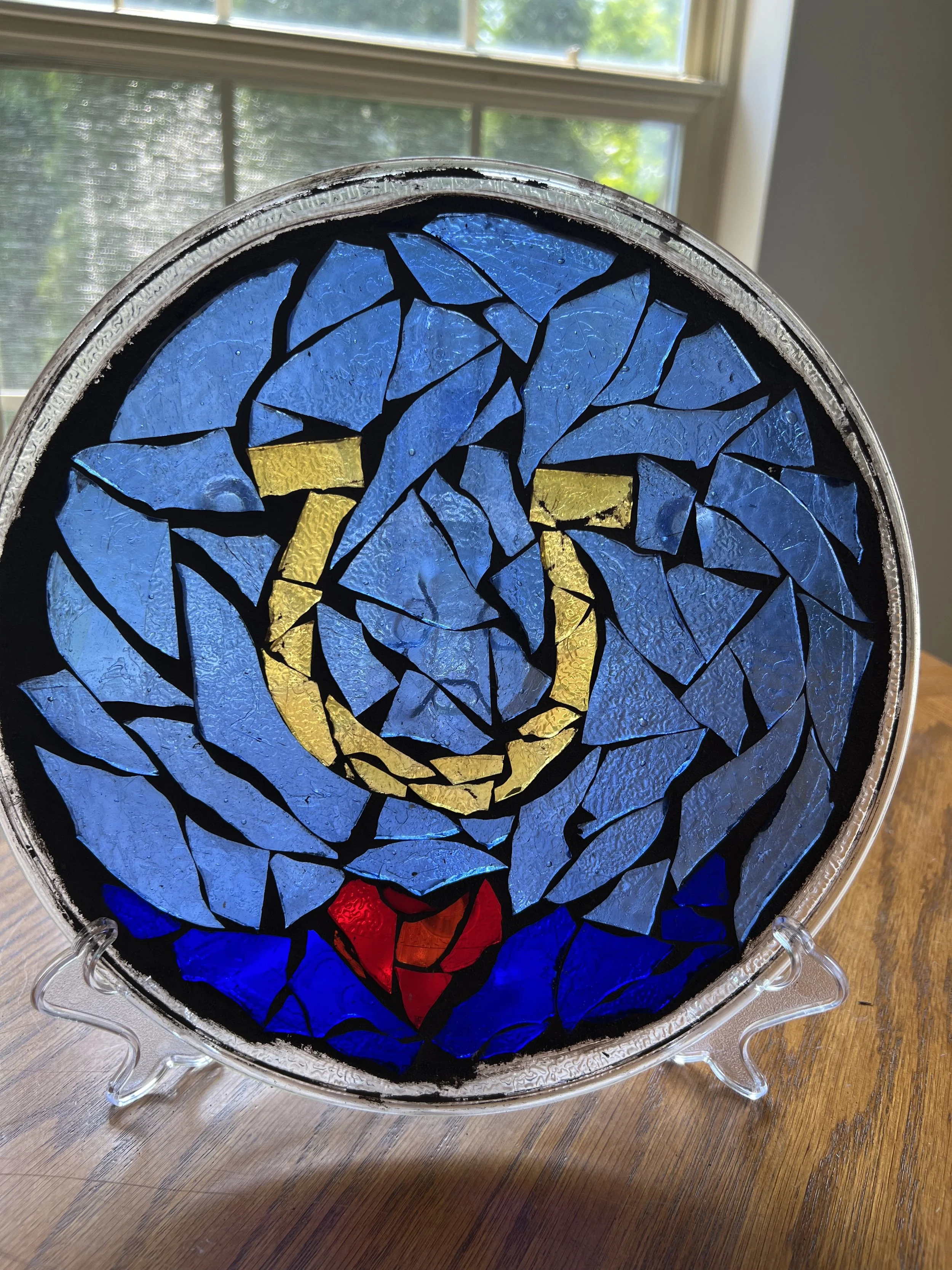 A stained glass art piece in a circular shape, depicting a dove with an olive branch in its beak, using blue, yellow, red, and black glass pieces.