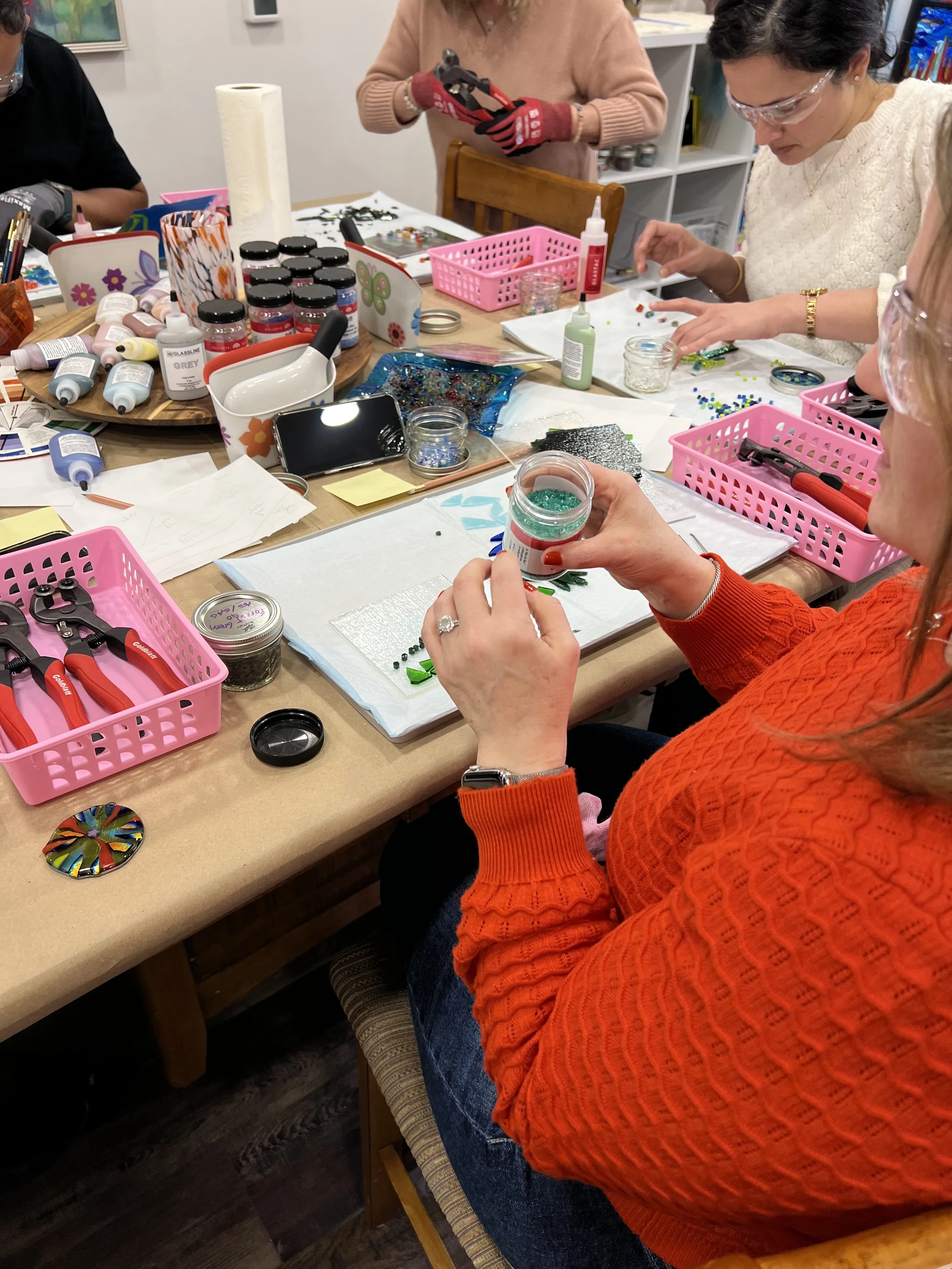 Women sitting at a table creating mosaic art with glass and beads, surrounded by jewelry-making supplies, tools, and organized containers of beads and glue.