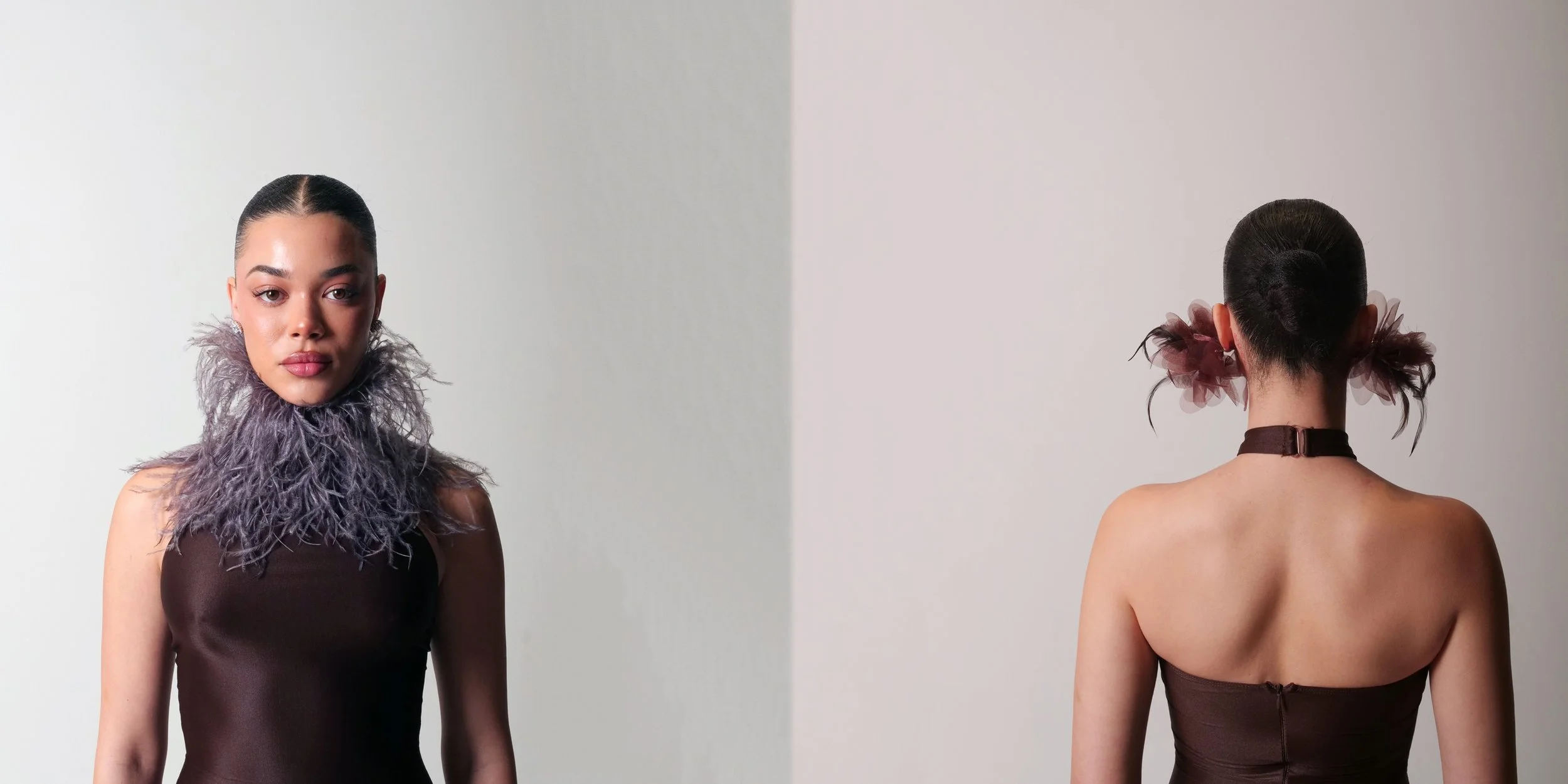 A woman with dark hair in a tight bun, wearing a black strapless dress, and a feathered collar around her neck. She stands facing forward with a neutral expression, with a plain light-colored background.