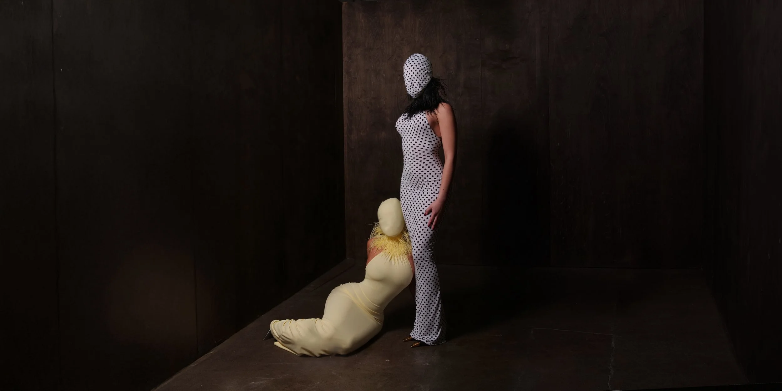 A woman in a polka dot dress and head covering stands in a dark room, looking at a figure dressed in a yellow costume with fuzzy collar that is kneeling and resting its head on her leg.