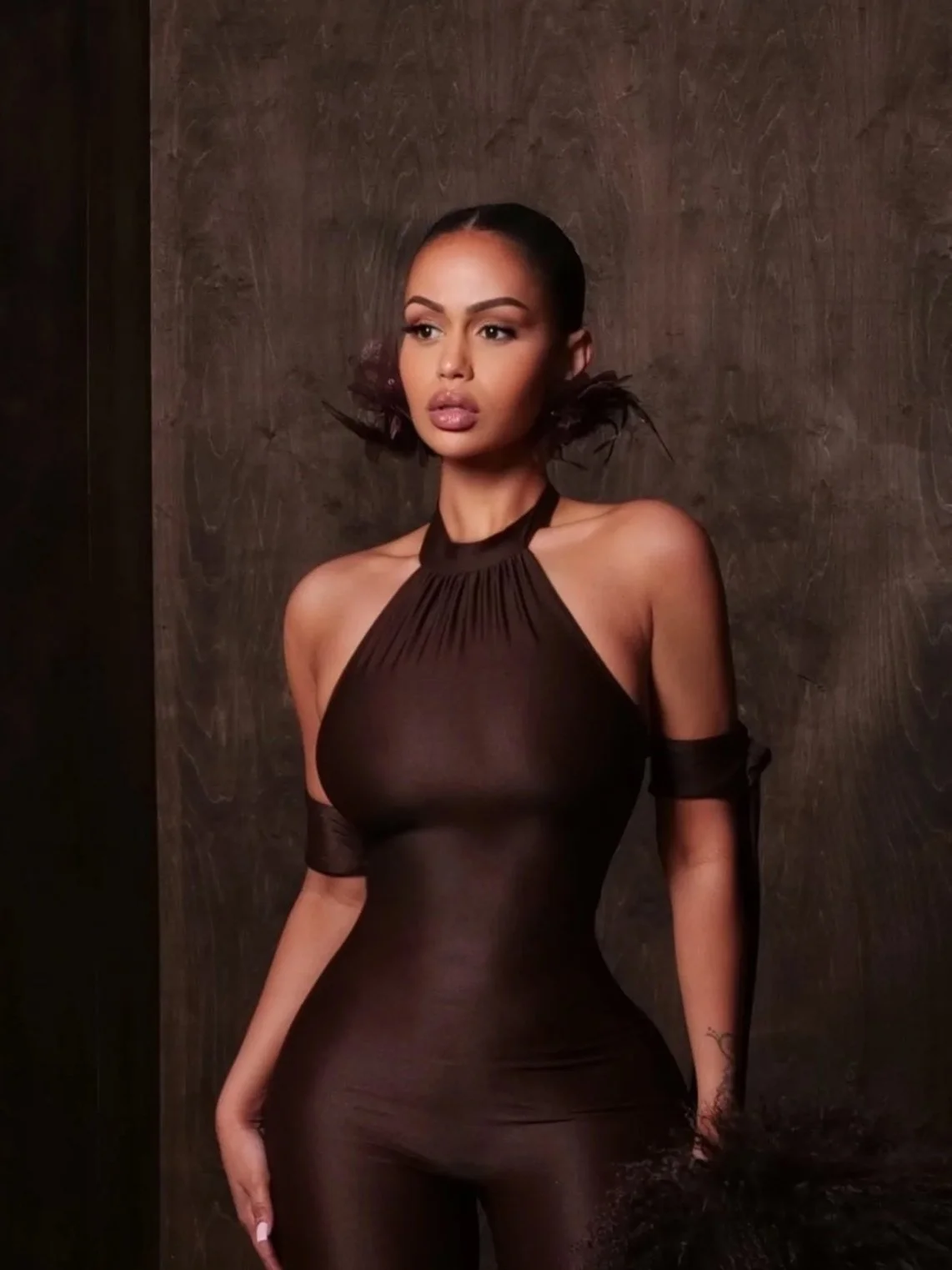 A woman with dark hair styled in a bob, wearing a sheer black halter dress with long sleeves, standing against a textured dark brown background.