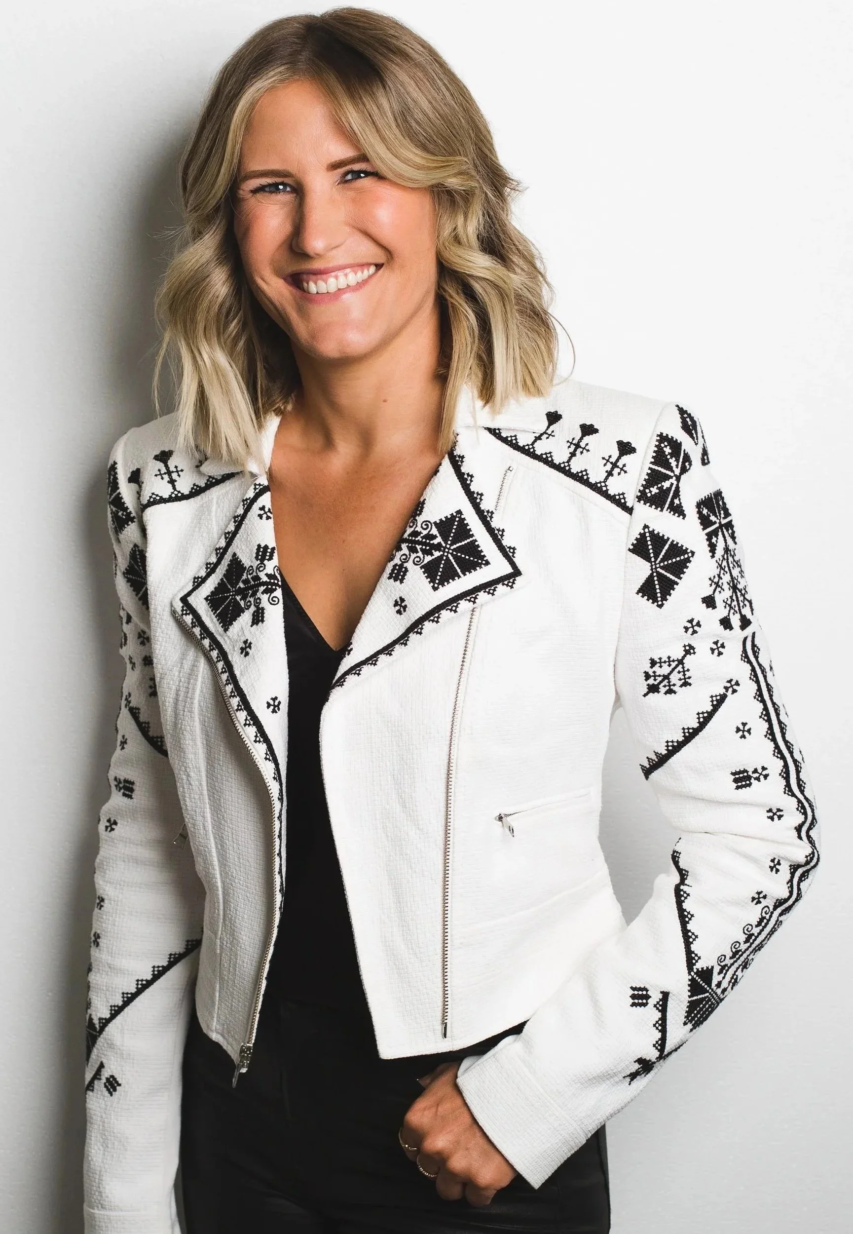 A woman with shoulder-length blonde hair, smiling, wearing a white jacket with black embroidery and a black top underneath, posed against a white wall.