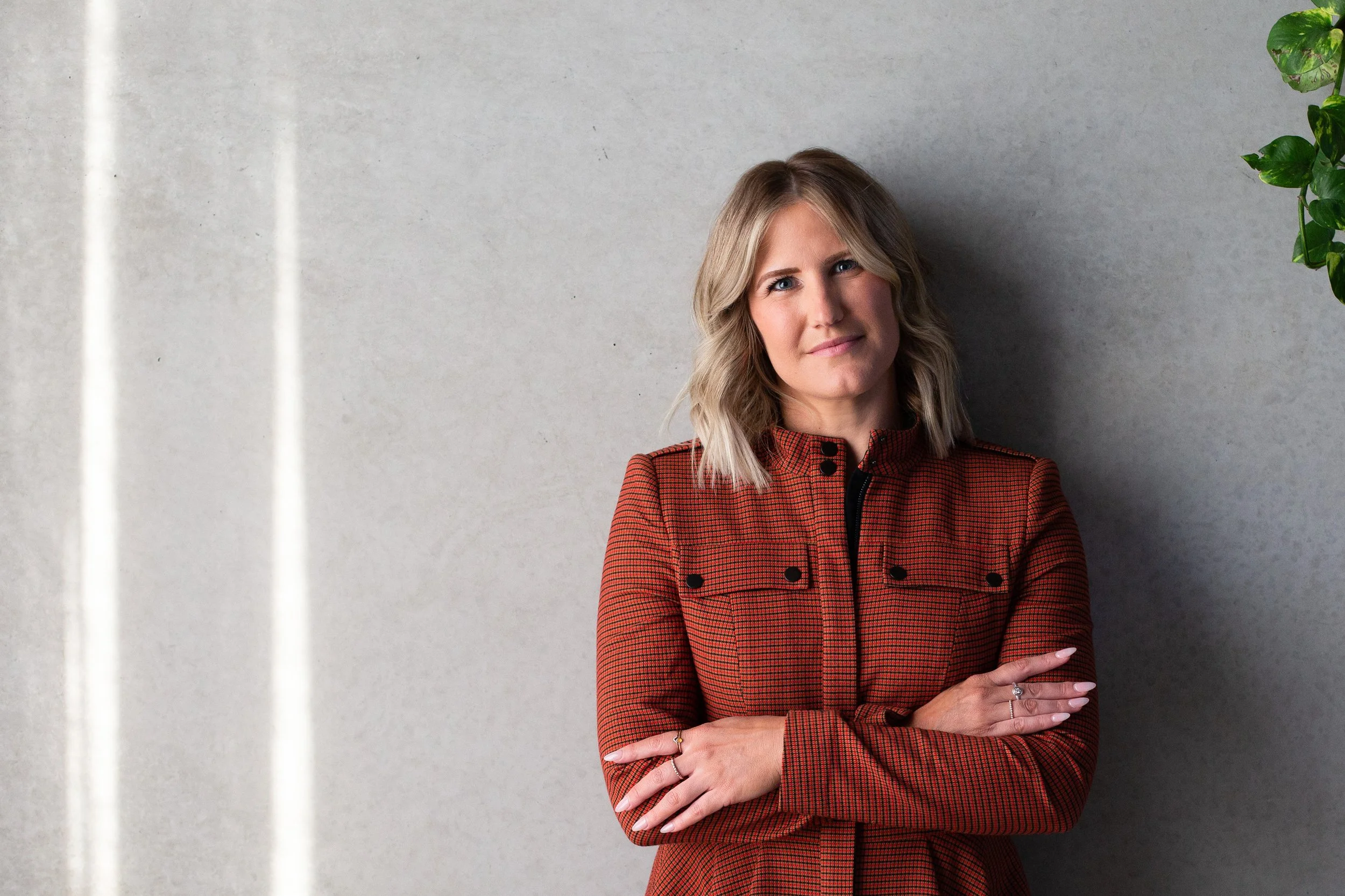 A woman with blonde hair in a red and black checkered jacket standing against a plain gray wall with a house plant on the right side.