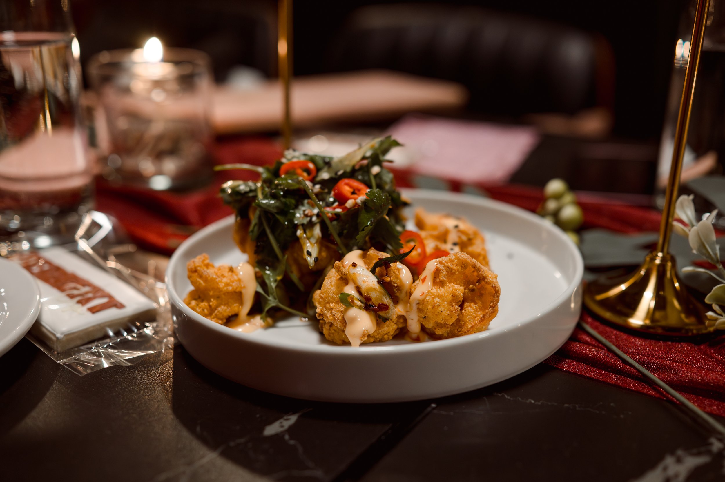 Fried chicken bites topped with a spicy mayonnaise sauce, garnished with fresh greens and sliced red chili, served on a white plate on a table with a red tablecloth.