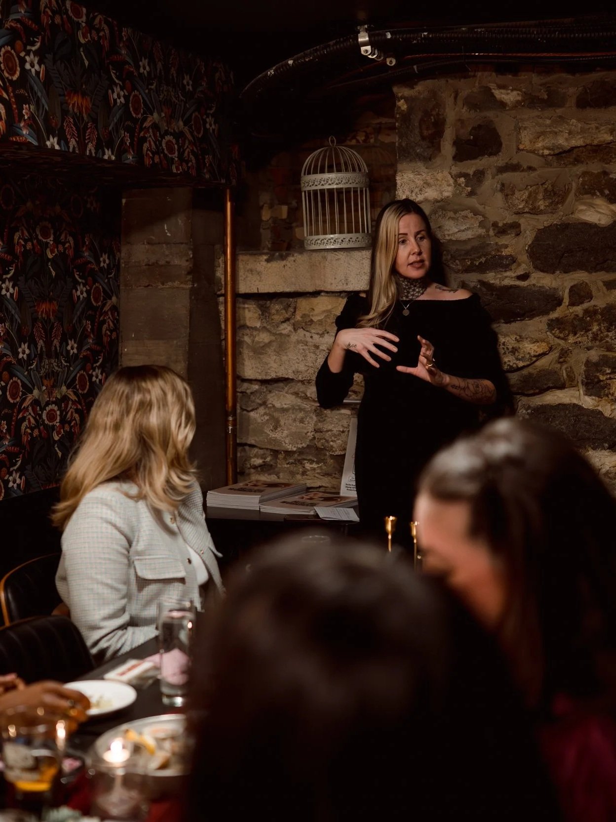Woman giving a speech or presentation in a dimly lit room with stone wall background, seated audience members visible in the foreground.
