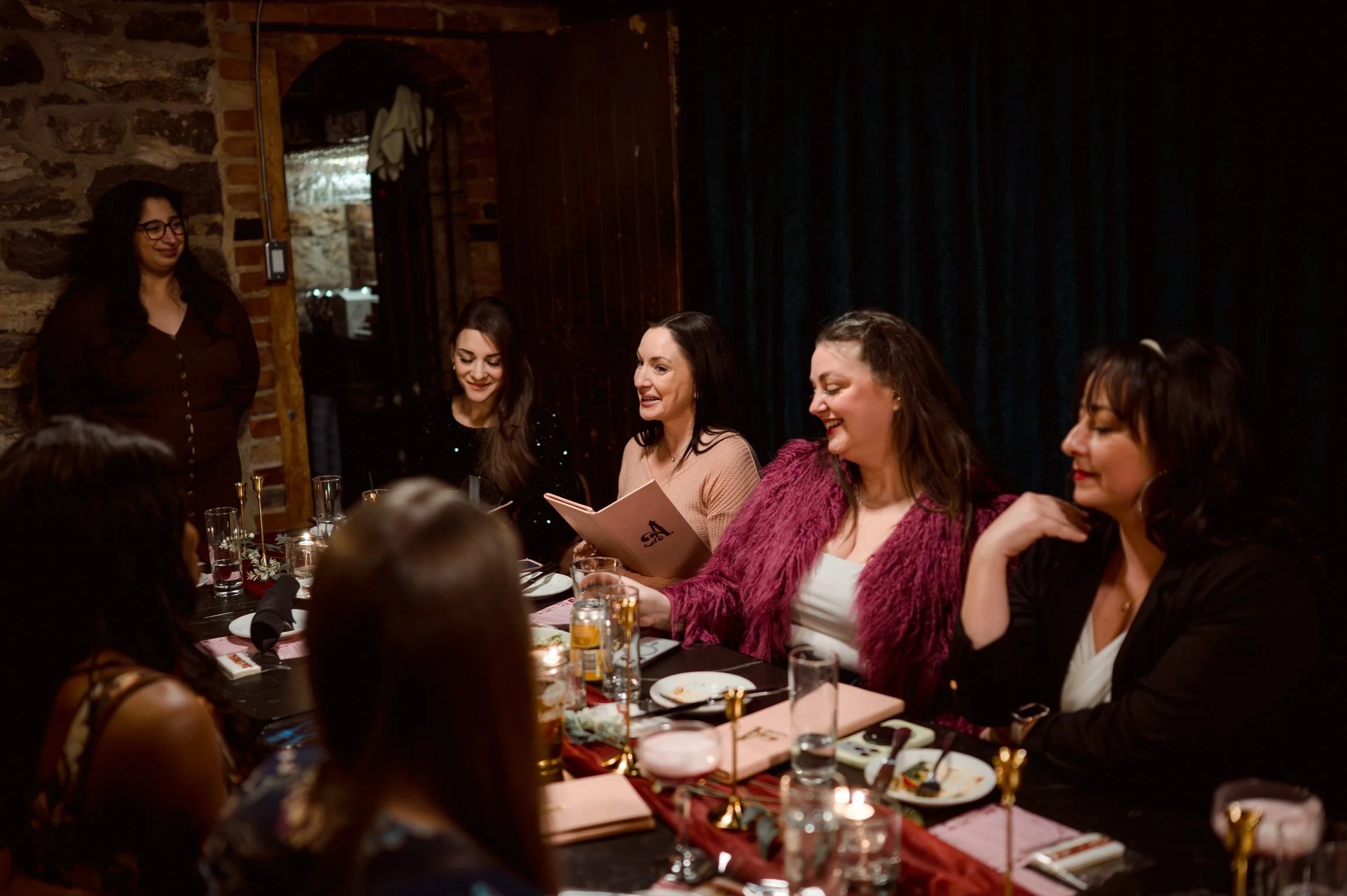Women gathered around a dining table, smiling and enjoying a celebration in a rustic setting with brick and wooden walls.