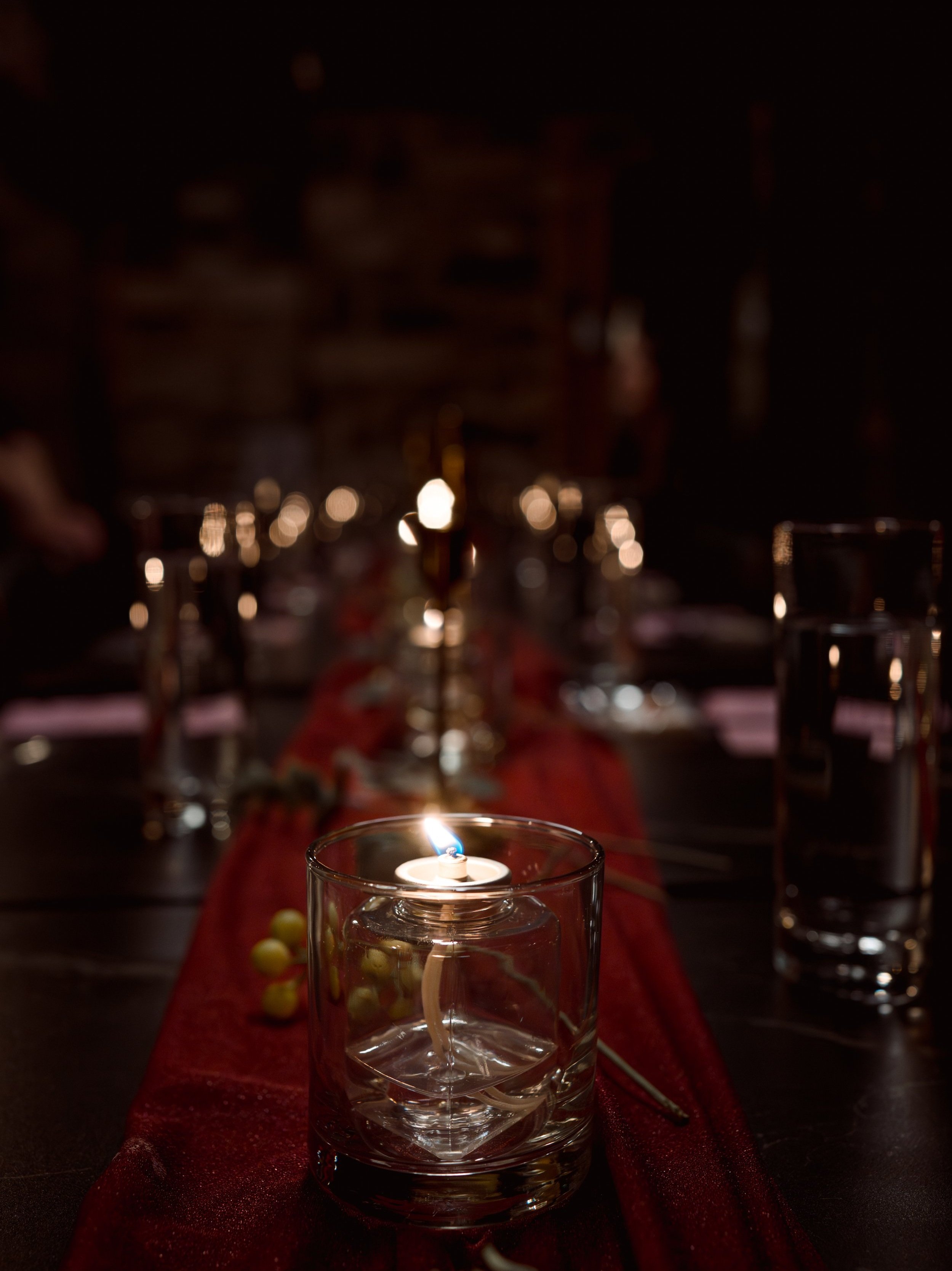 A lit white candle inside a clear glass holder on a dark table, with a red table runner and multiple empty glasses, in a dimly-lit indoor setting.