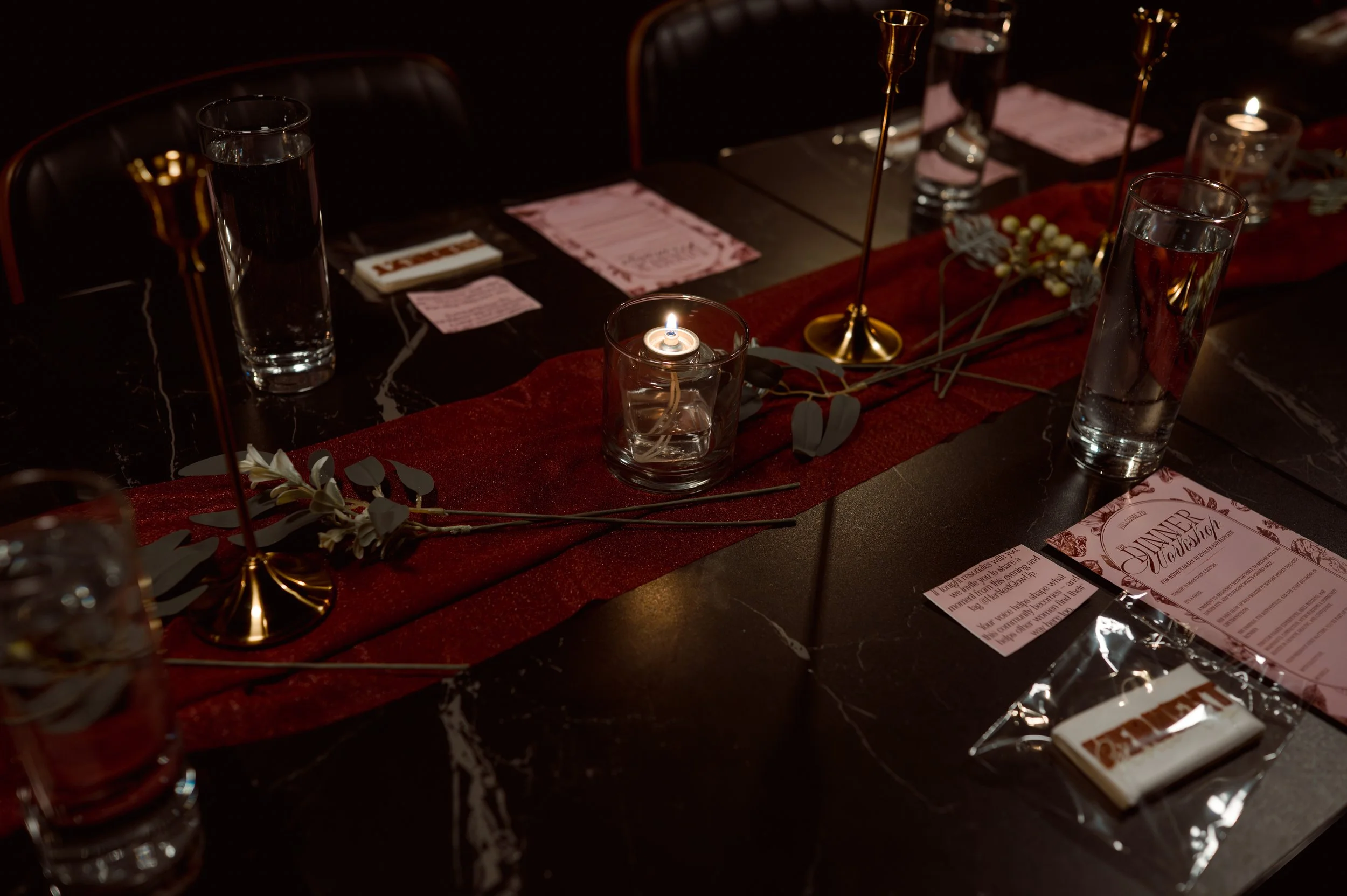 A dark dining table decorated with a red runner, candles, green leaves, and place settings, including pink menus, glasses of water, and small wrapped chocolates.