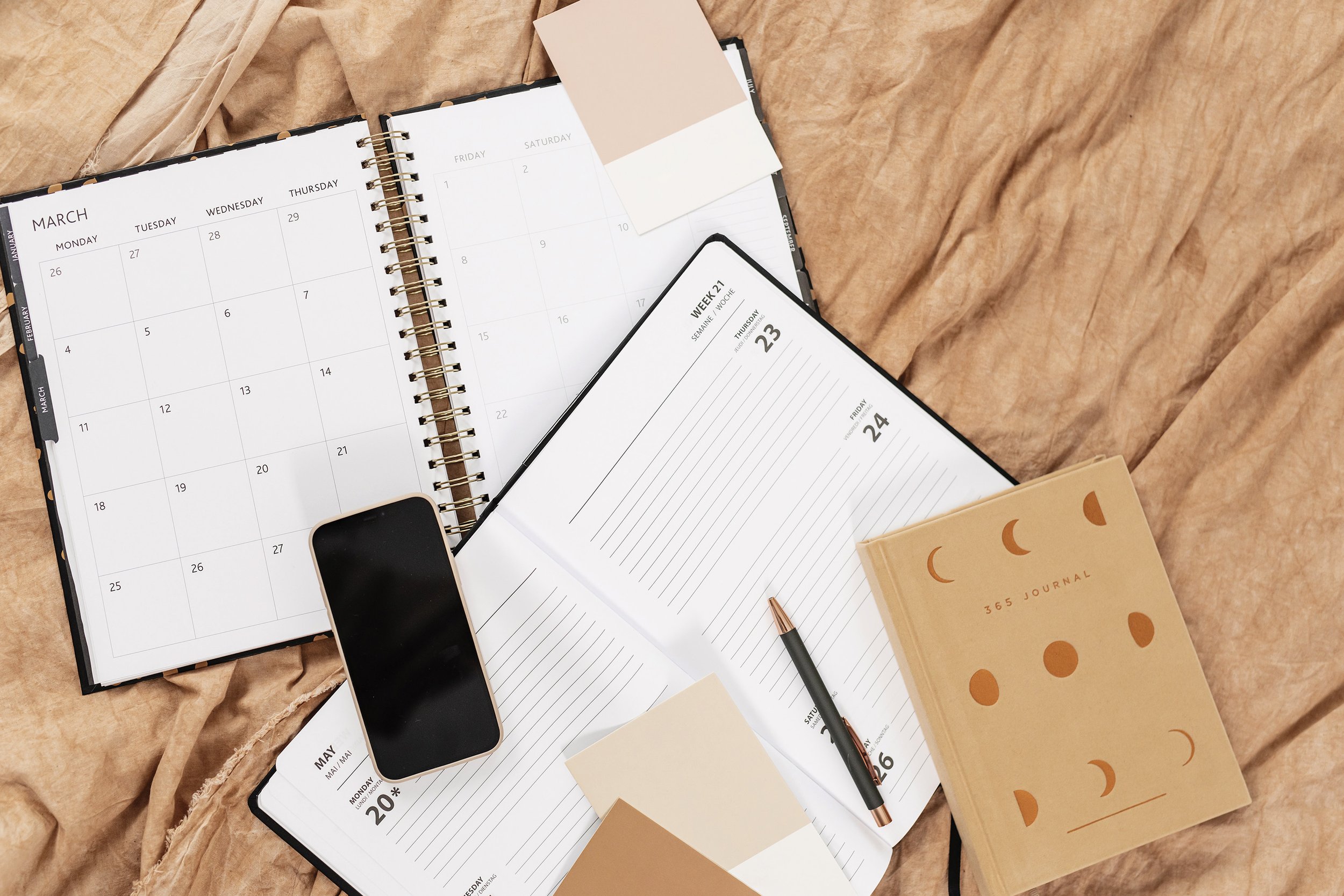 Open planner with calendar pages for March, April, and May, a black smartphone with a blank screen, a black and rose gold pen, a beige journal with moon phases on the cover, and a few sticky notes on crinkled brown paper background.