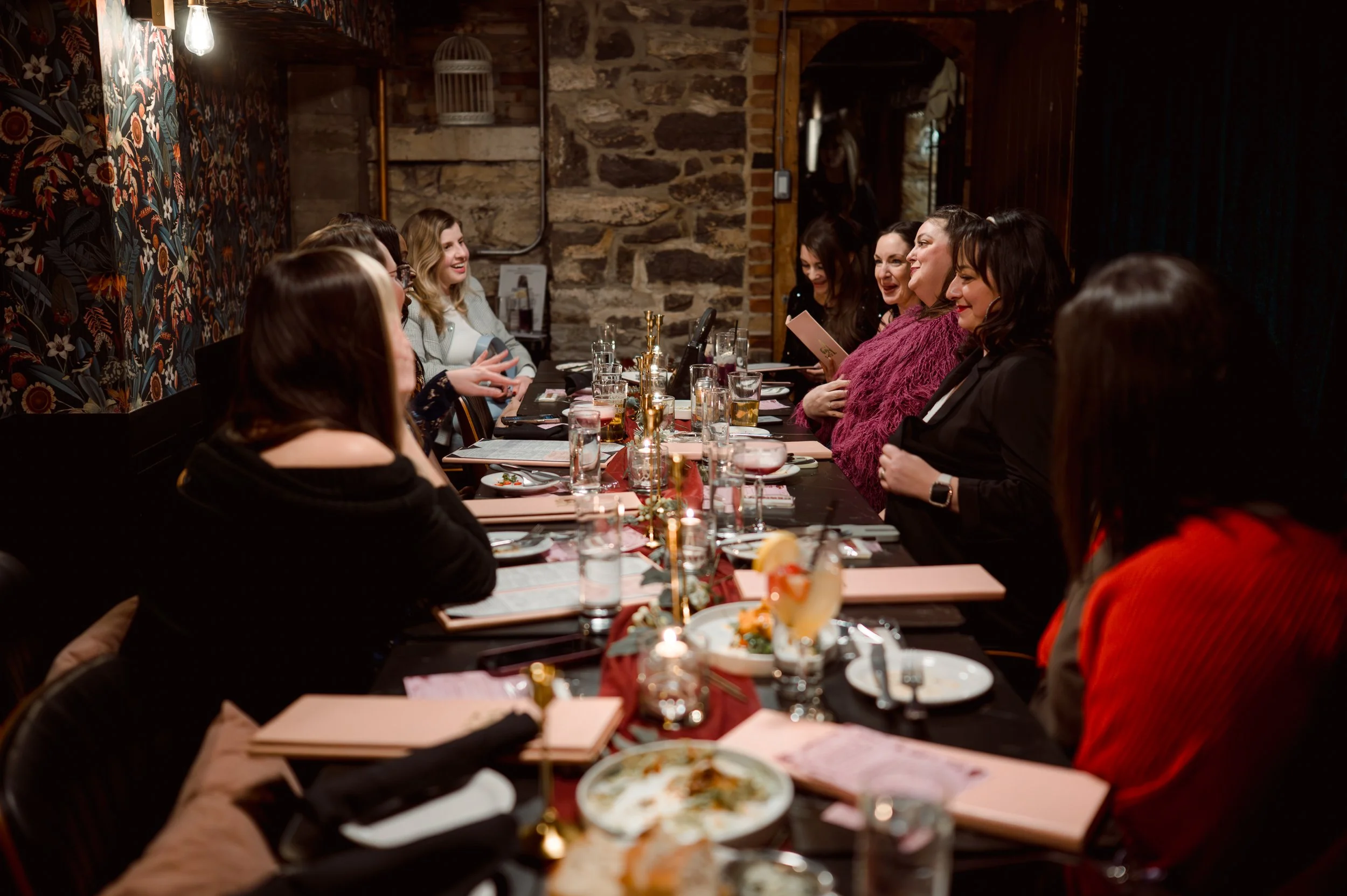 Women sitting around a long table in a cozy restaurant, engaged in conversation, with food and drinks on the table.