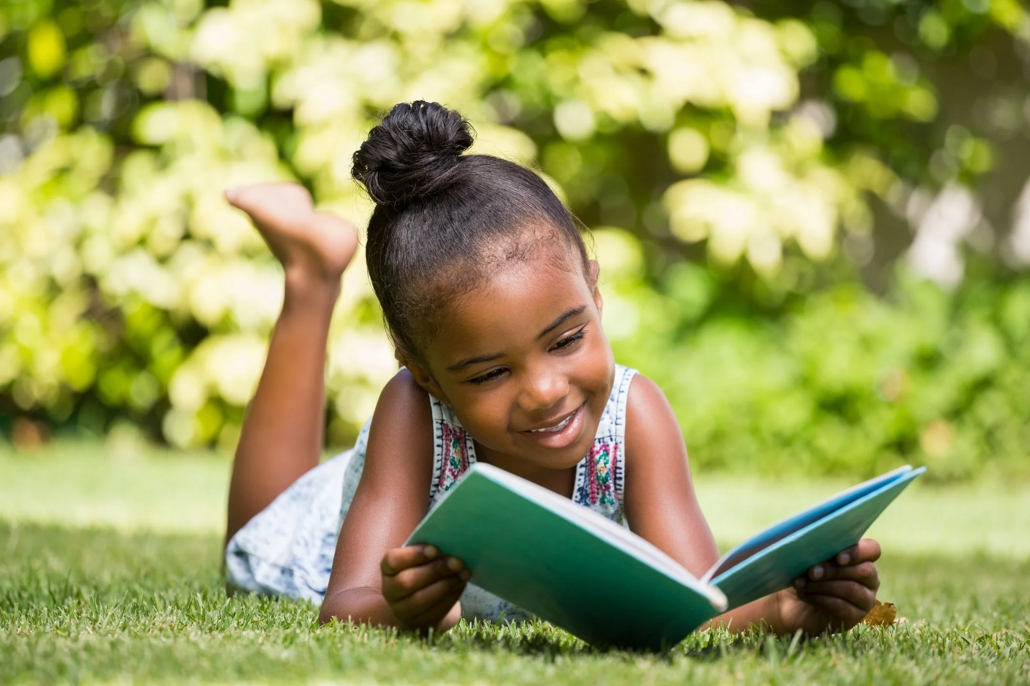 Young girl with dark hair in a bun lying on grass, reading a book outdoors with green trees in the background.