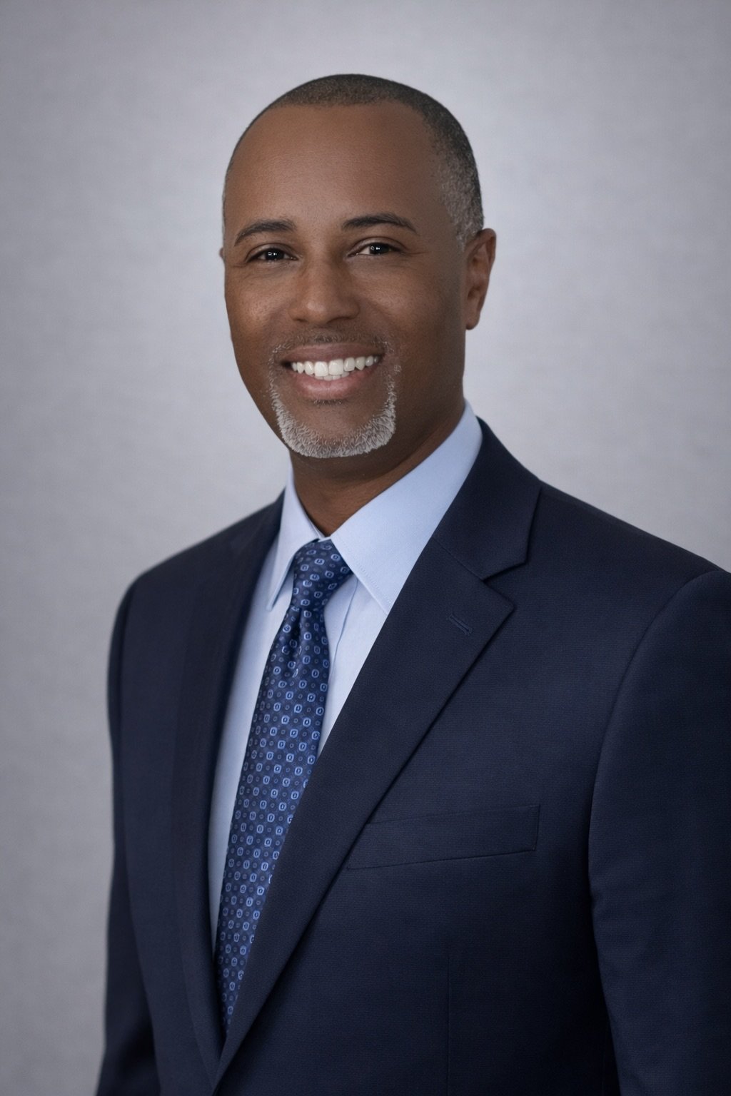 A professional headshot of an African American man wearing a dark suit, light blue shirt, and a patterned tie, smiling at the camera against a neutral background.