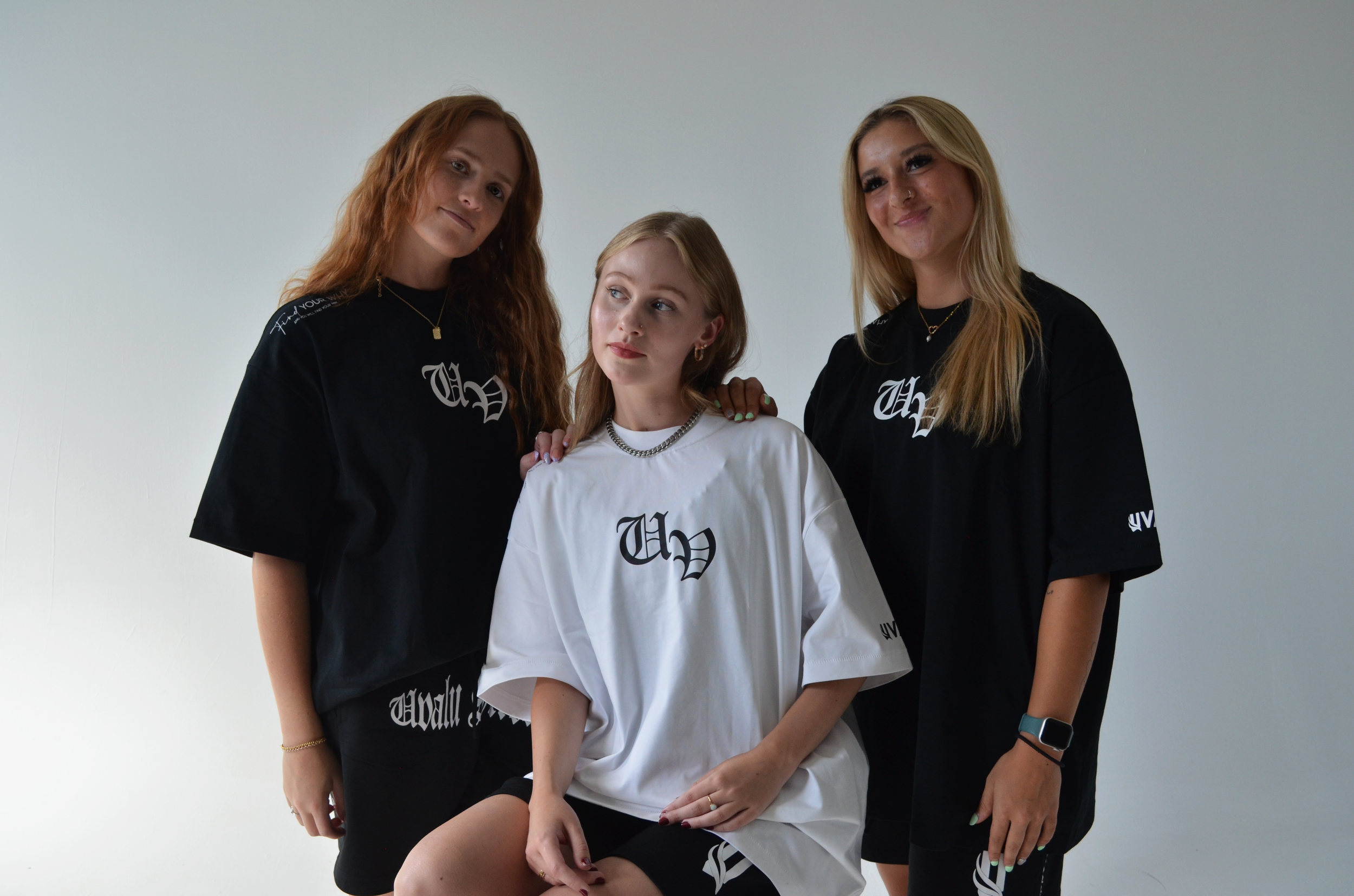 Three young women posing together against a plain white background, wearing matching black and white oversized T-shirts with a gothic-style logo.